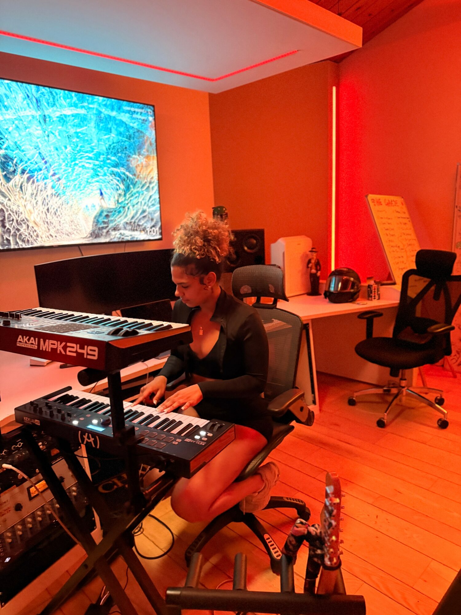 Person with curly hair playing keyboard in a room with red walls and a large screen, wooden floor, and office chairs.