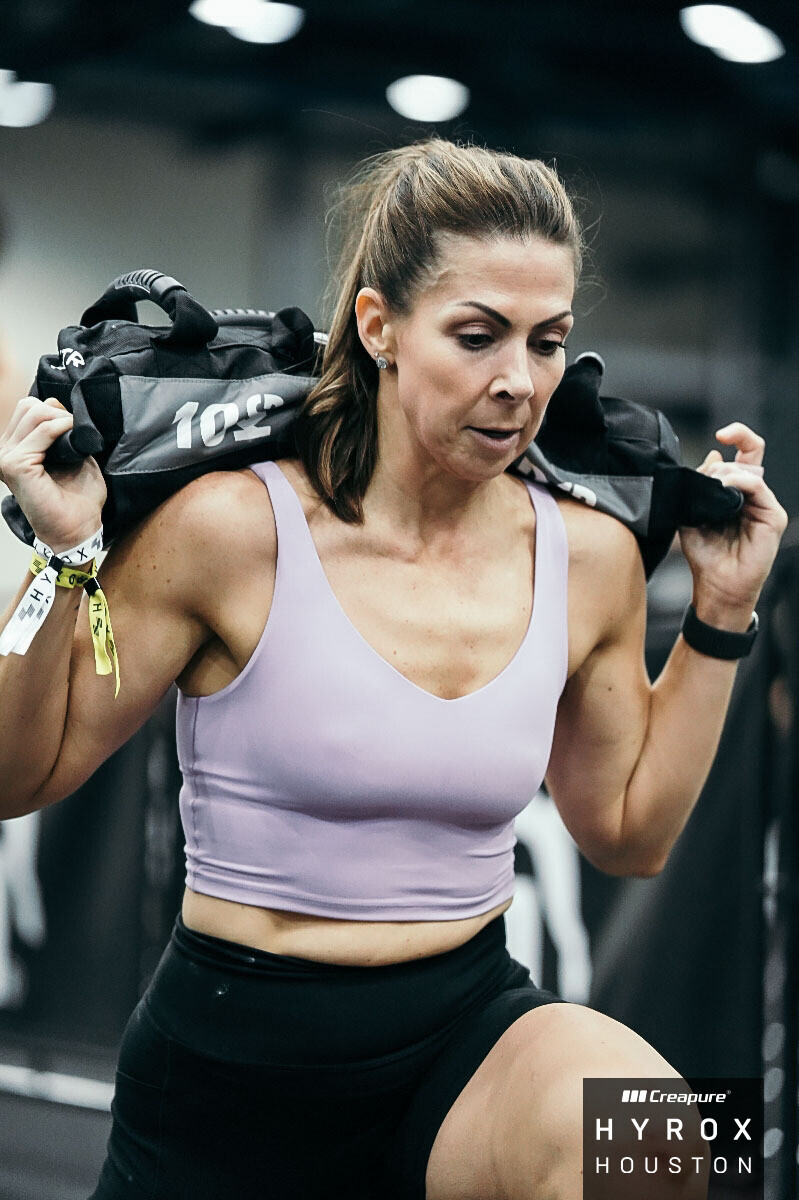 Woman in workout clothes lifting a weighted bag on her shoulders in a gym, focused expression, dark background.