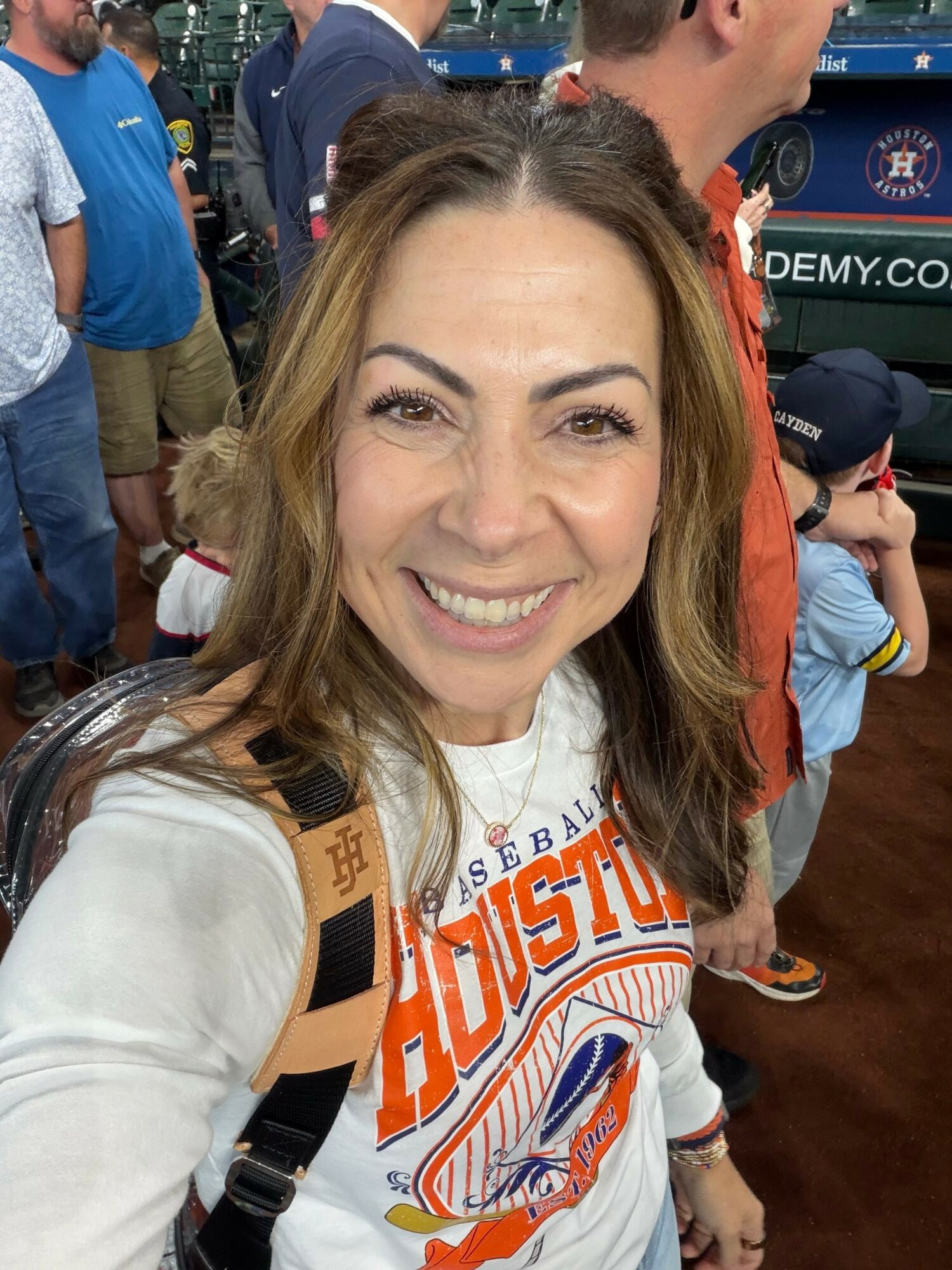 Smiling woman with shoulder-length hair in a white Houston Astros shirt at a baseball game, crowd in background.
