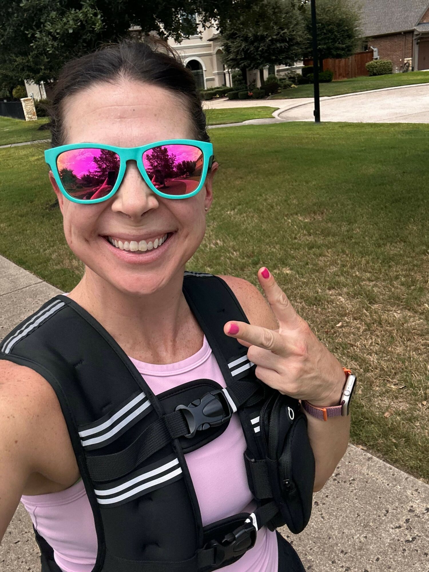 Smiling woman wearing sunglasses and a pink top, pointing with her finger, outdoors in a park-like setting.