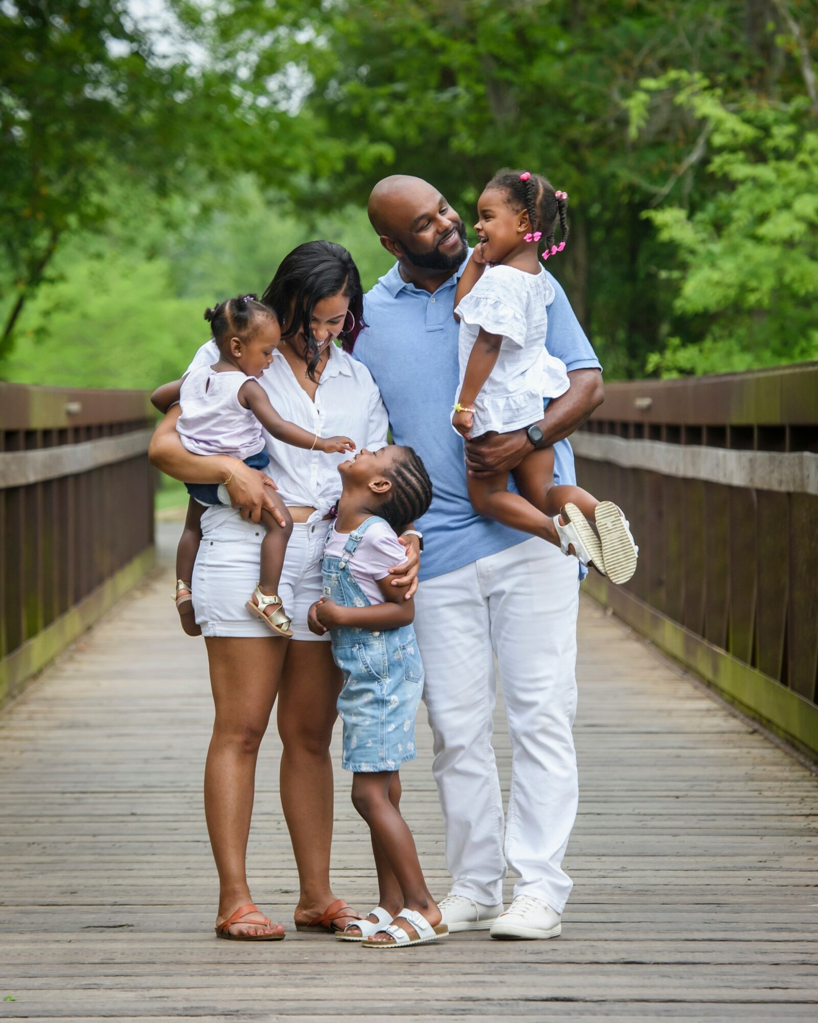 Family of six standing on a wooden bridge outdoors, smiling and interacting with each other, surrounded by green trees.