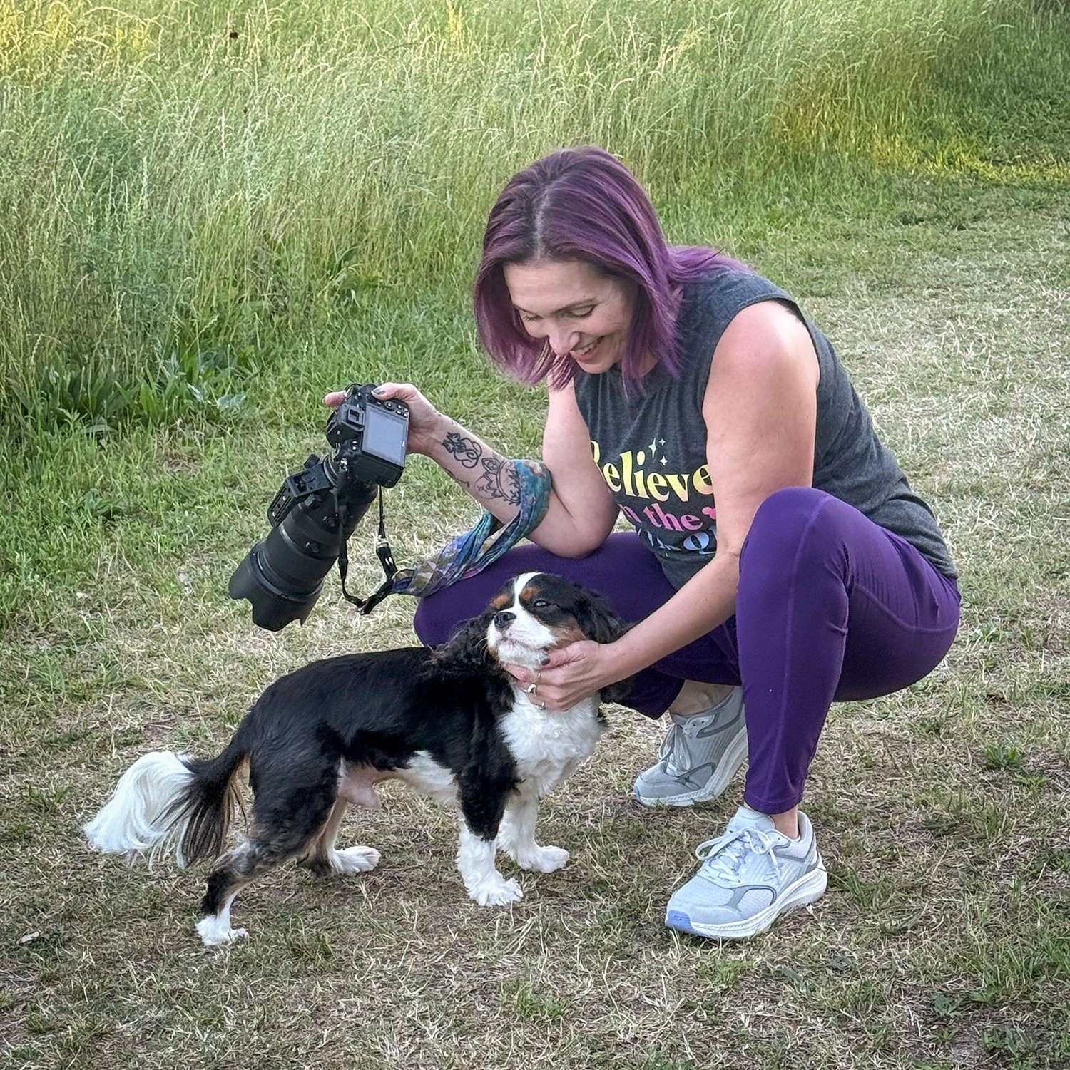 Woman with purple hair smiling, holding a camera in one hand and petting a small black and white dog outdoors.