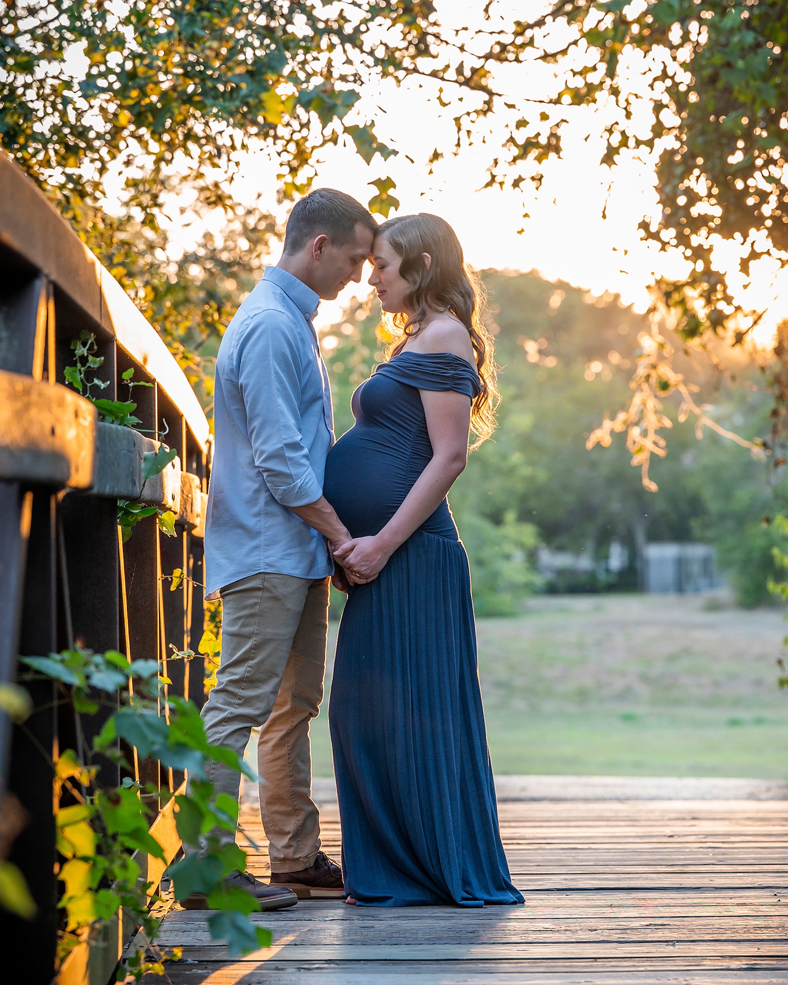 Pregnant woman and man holding hands on a bridge at sunset, trees in background, warm lighting, outdoor setting.