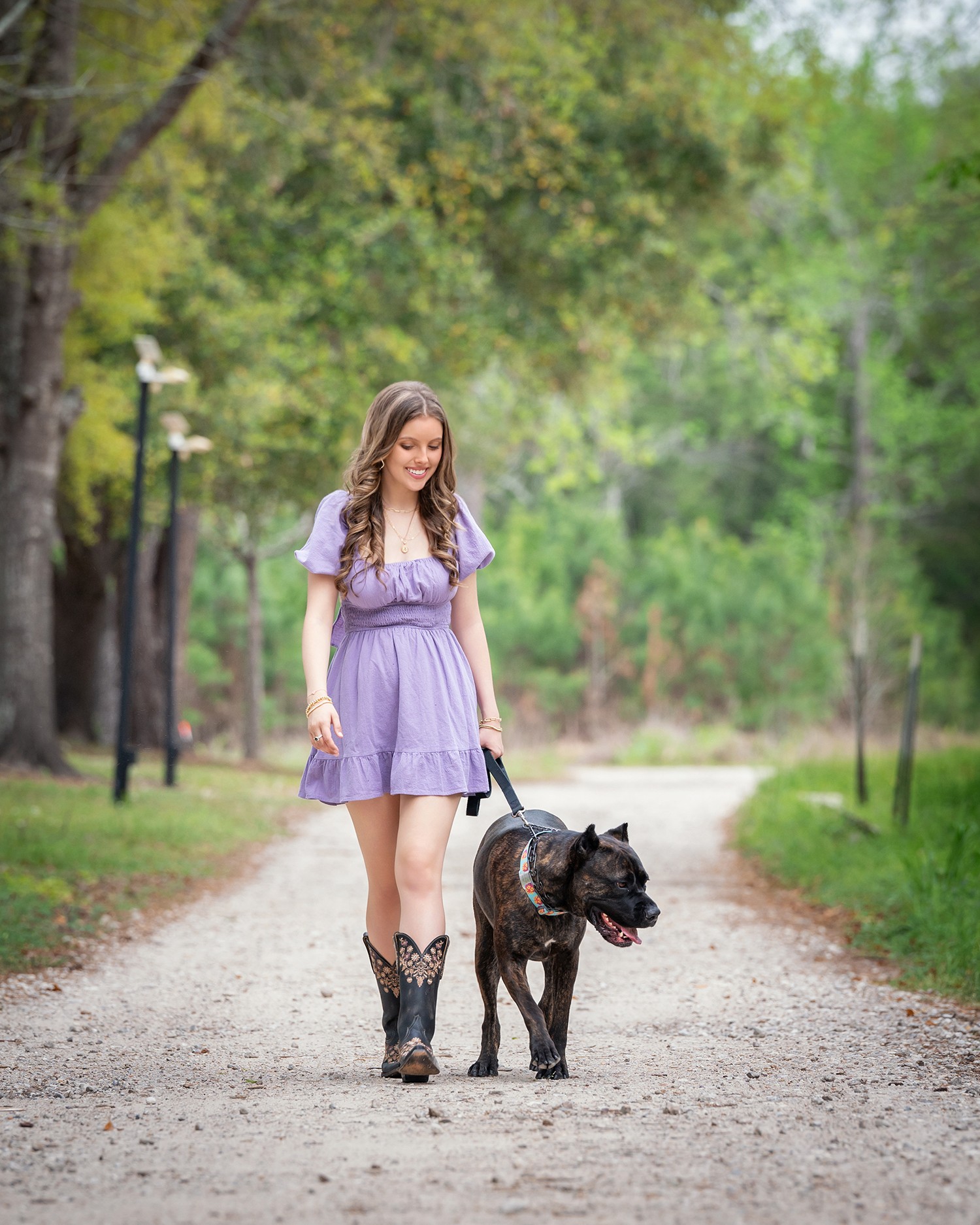 Young woman walking a dog on a park path surrounded by green trees, wearing a purple dress and cowboy boots.