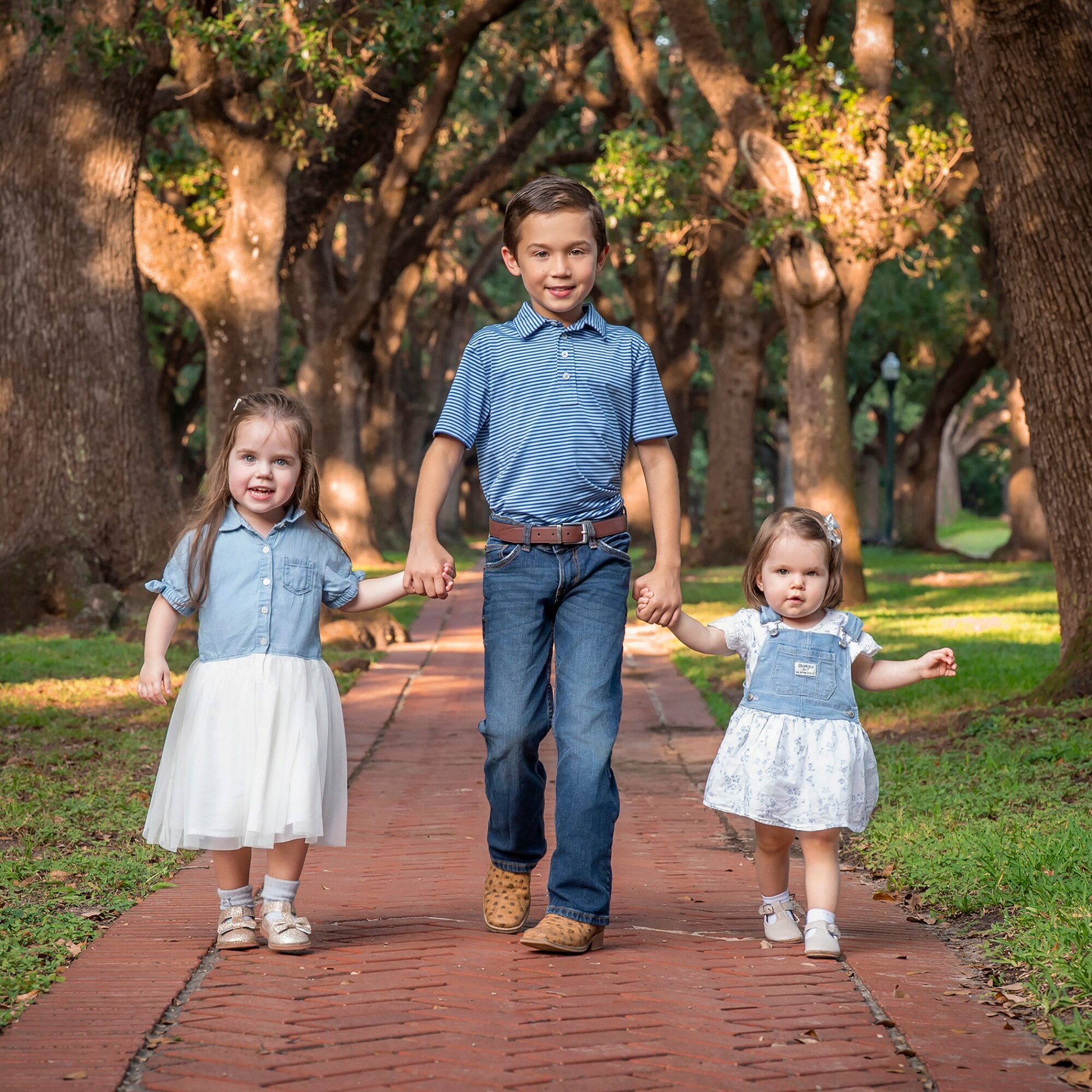 Three children walking hand in hand on a brick path in a park with large trees and green grass.