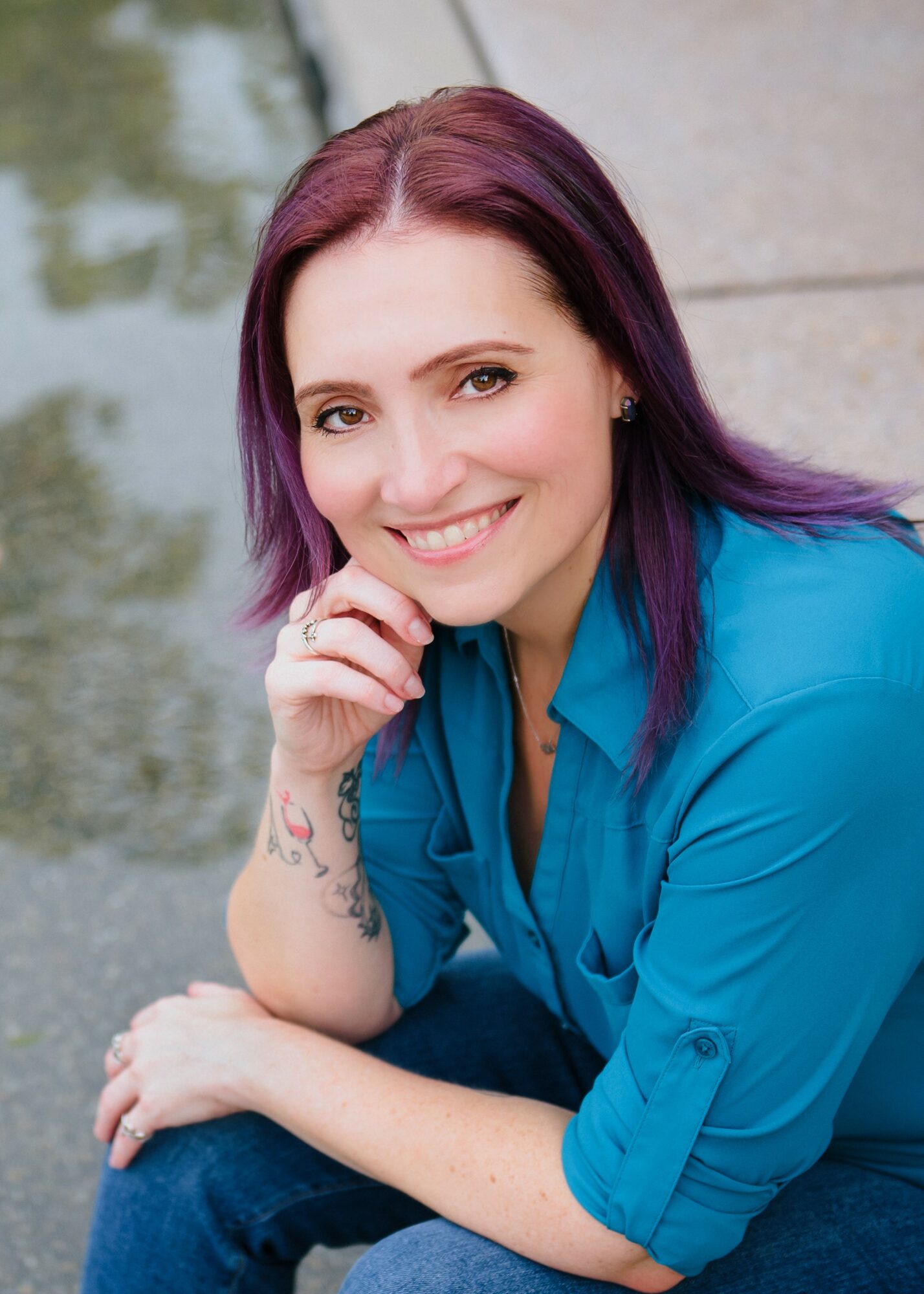 Smiling woman with purple hair wearing a blue shirt, sitting outdoors near water, resting her chin on her hand.