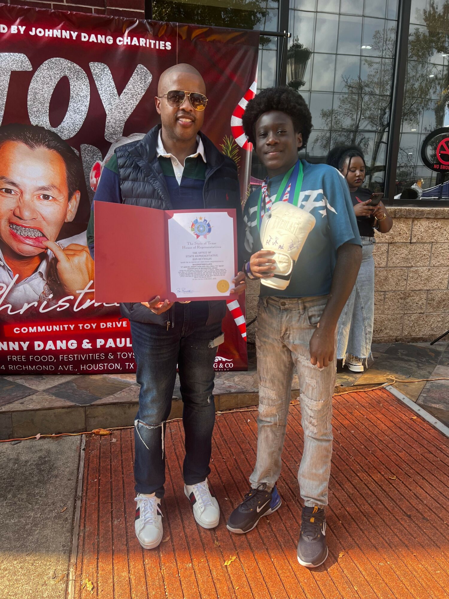 Two people standing outdoors, one holding a certificate, with a red banner and glass building in background.