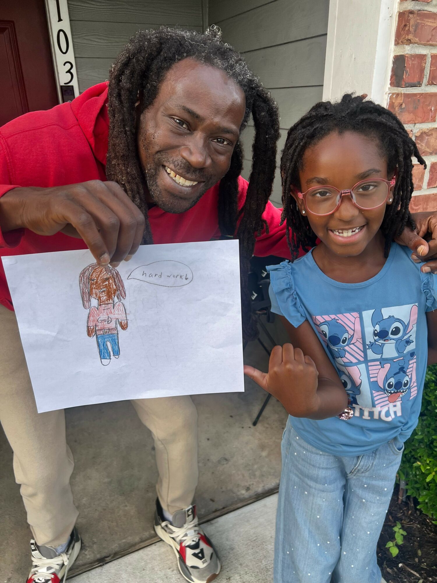Two smiling people, one holding a drawing of a person, standing outside near a brick wall and greenery.