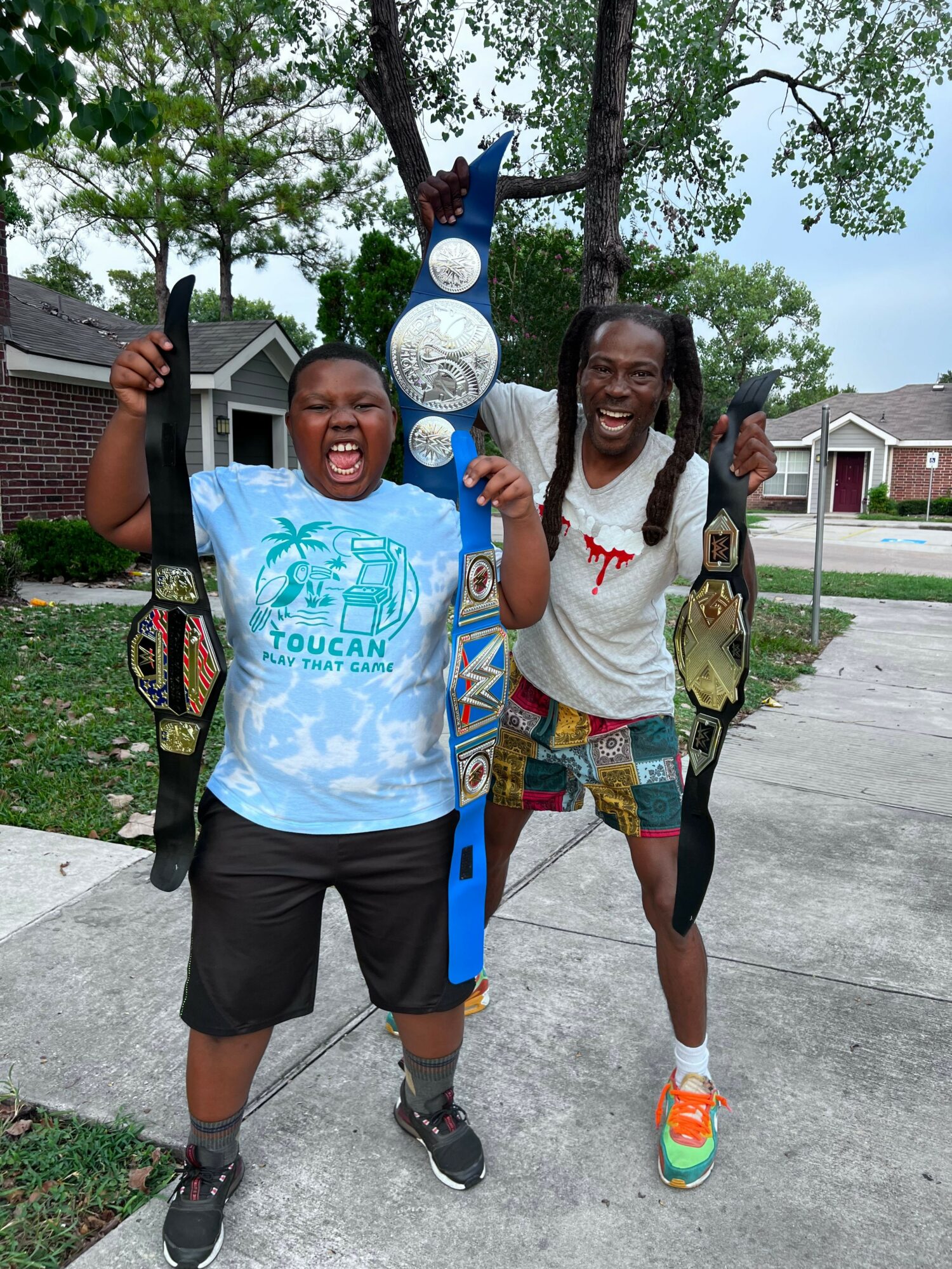 Two smiling women standing outdoors on a sidewalk, holding drinks and giving thumbs-up gestures, with trees and houses in background.