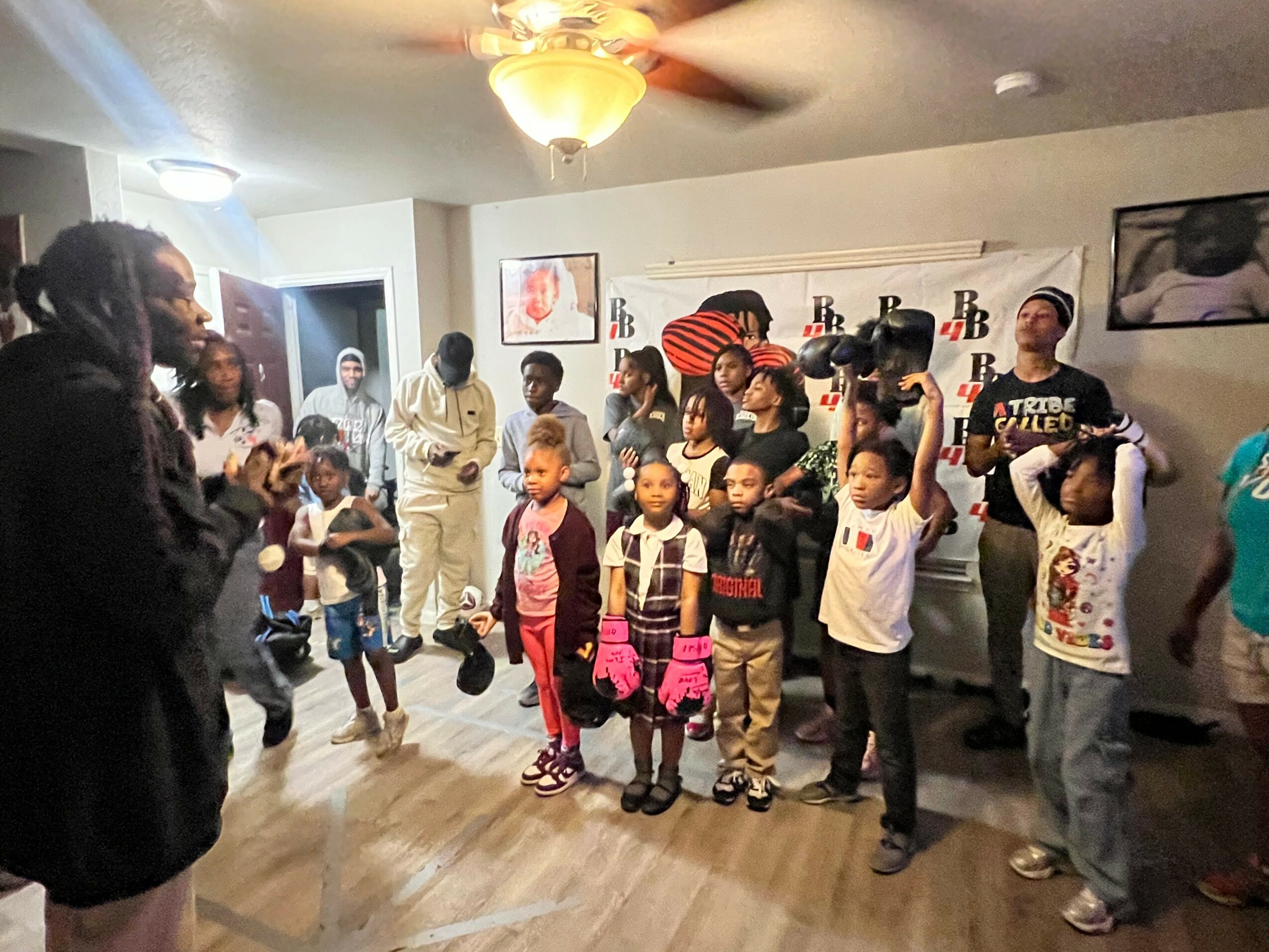 Group of children and adults gathered indoors, some children holding hats, in a room with a banner and wall decorations.