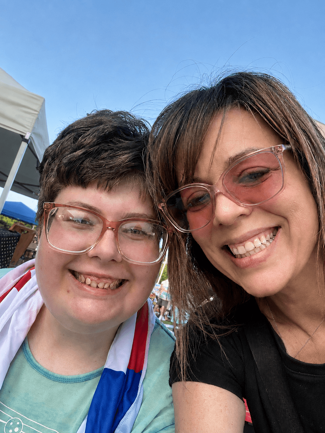 Two smiling women with glasses taking a selfie outdoors on a sunny day, with a tent in the background.