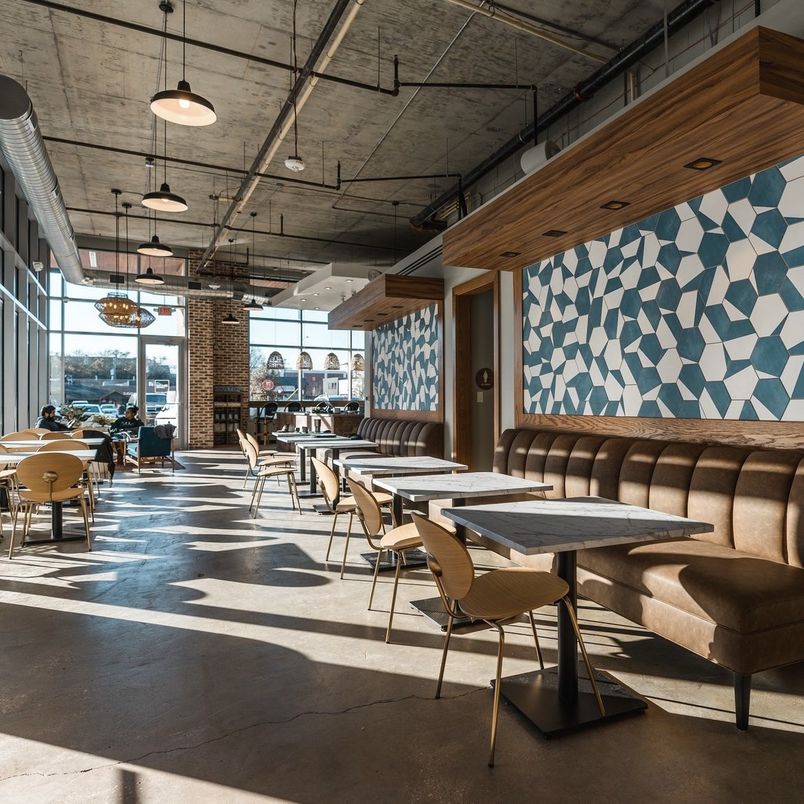 Interior of a modern cafe with tables, chairs, and a patterned blue wall, sunlight streaming through large windows, ceiling lights, and a brick pillar.