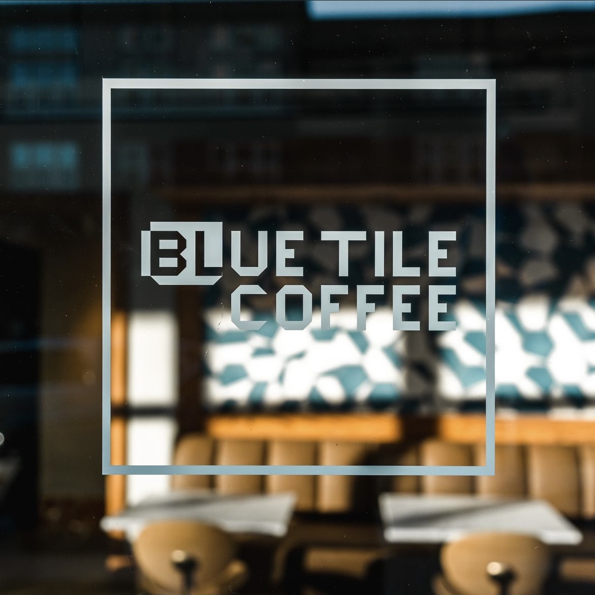 Sign with white text 'BLUE TILE COFFEE' inside a white square on a glass window, with blurred interior background.