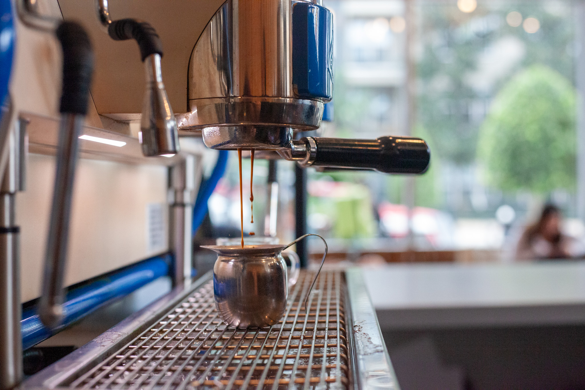 Close-up of a coffee machine pouring coffee into a cup on a metal grid, with a blurred window view outside.
