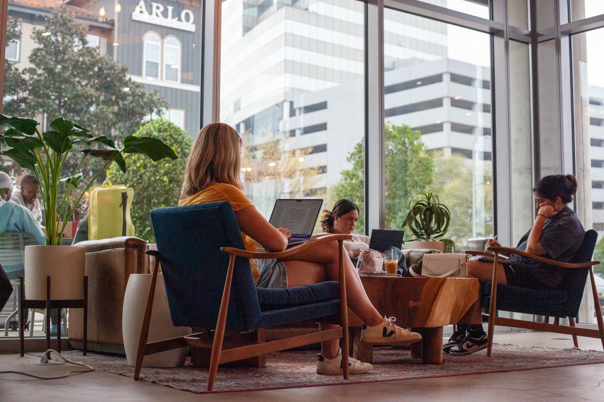 Two women sit in a modern cafe with large windows, working on laptops, with plants and city buildings outside.