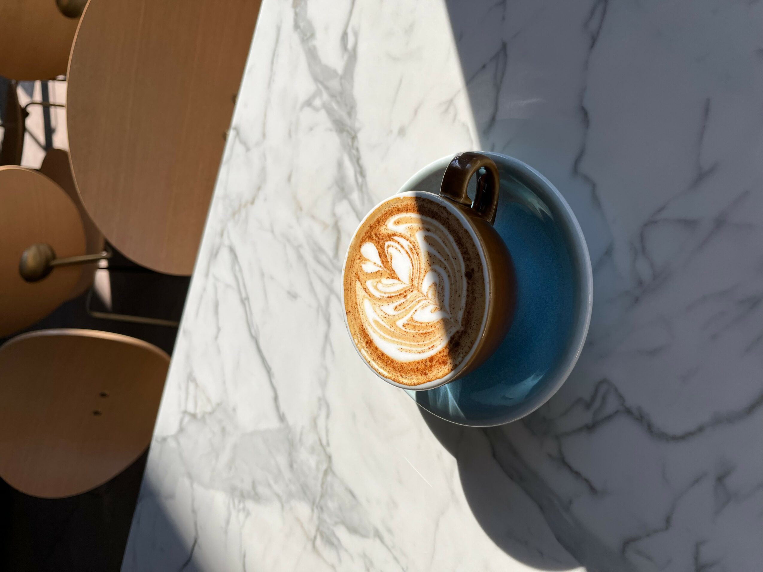 A cup of coffee with latte art on a marble table, sunlight casting shadows, in a cafe setting.