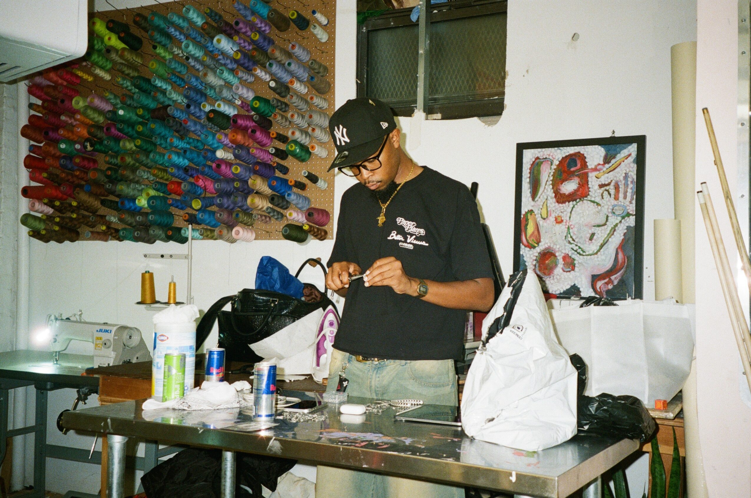 Young person wearing a cap and glasses, standing at a cluttered table with art supplies and bags, in a room with artwork on the wall.