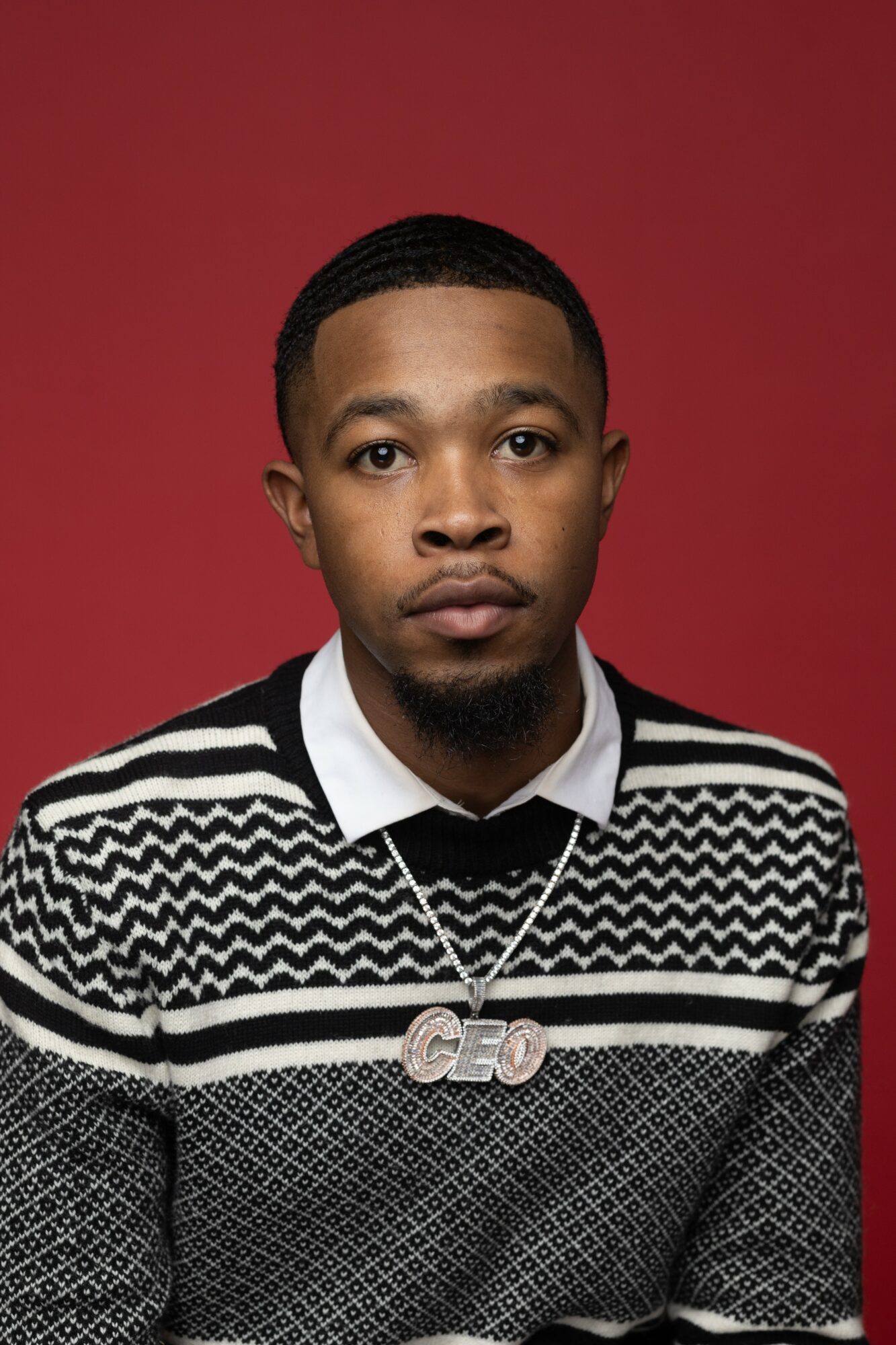 Young man with short hair and a goatee, wearing a patterned sweater and a chain necklace, against a red background.