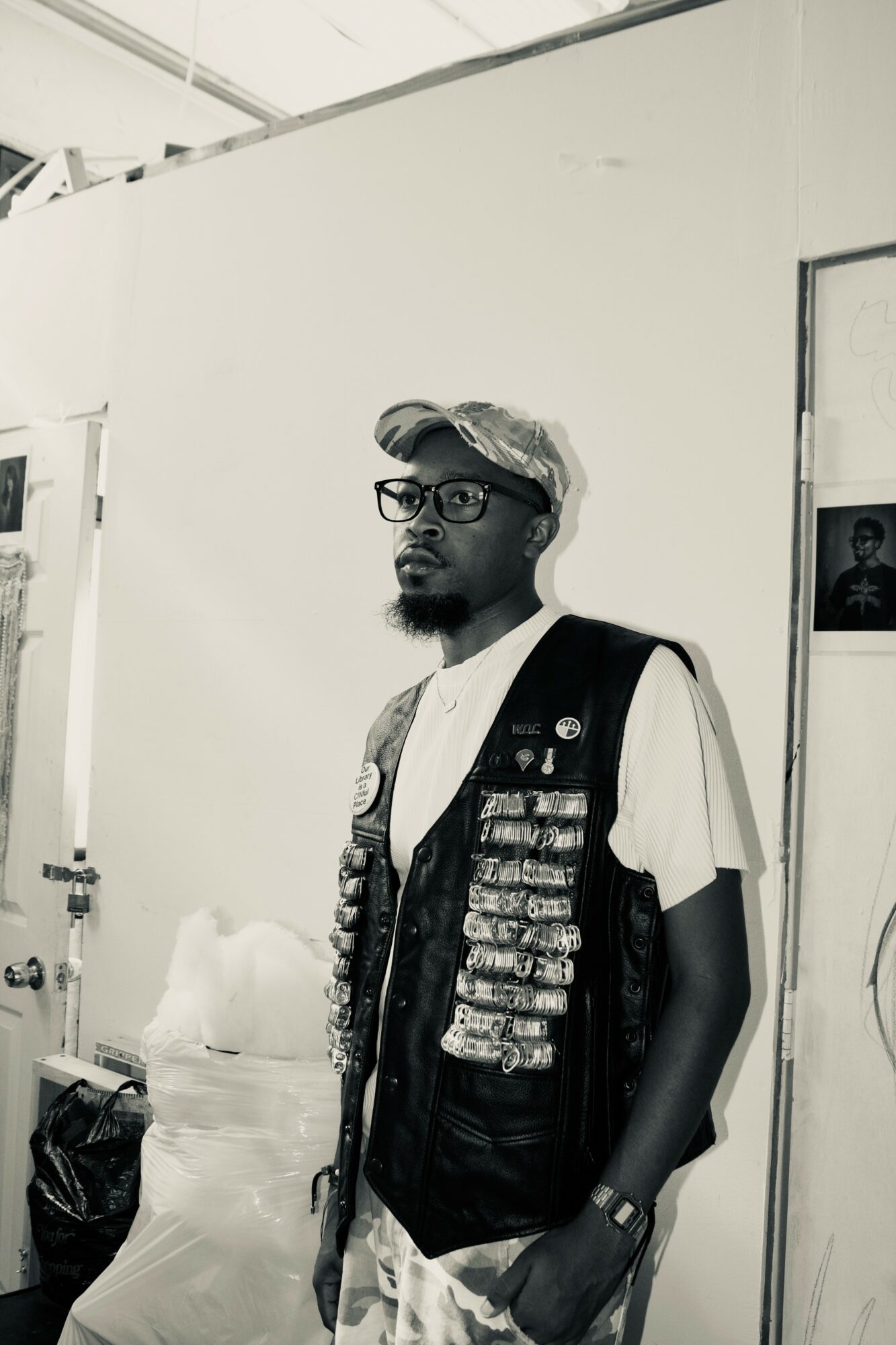 Man with glasses and a cap stands indoors, wearing a vest with patches over a white t-shirt, with a white wall behind.