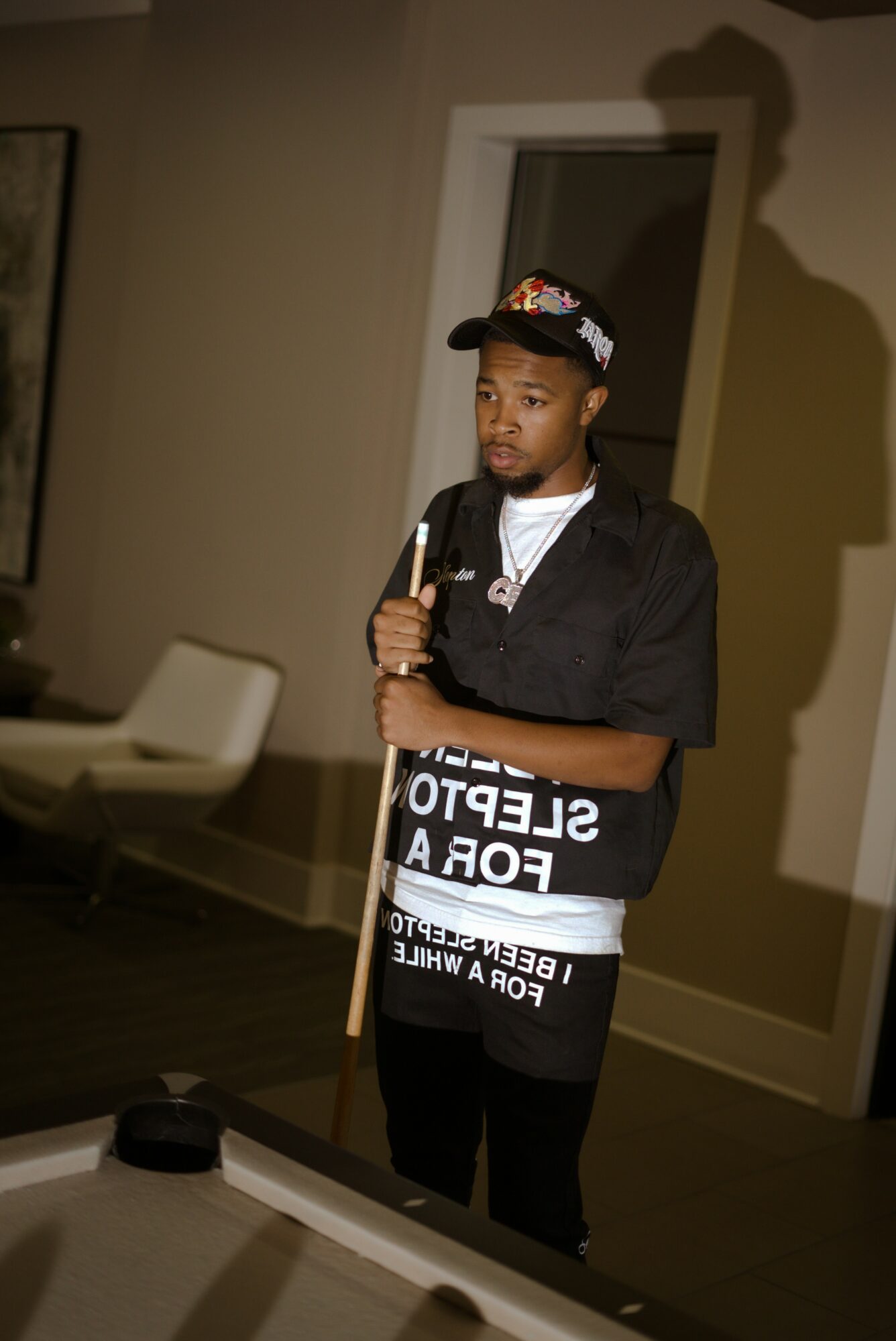 Young man in a cap and black shirt holding a pool cue in a room with beige walls and a window.