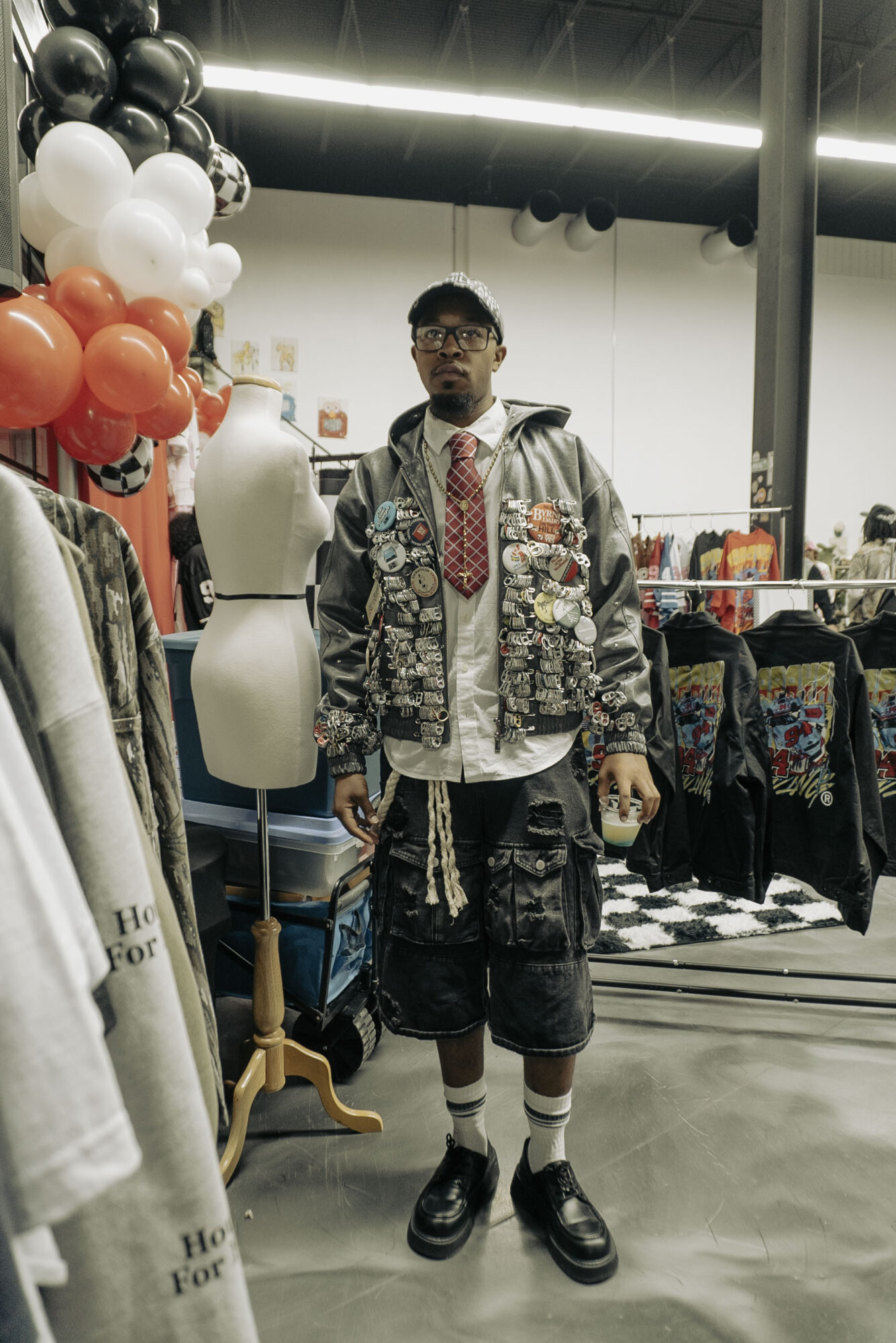 Young man with glasses and a patterned jacket standing in a store with balloons and clothing displays behind him.