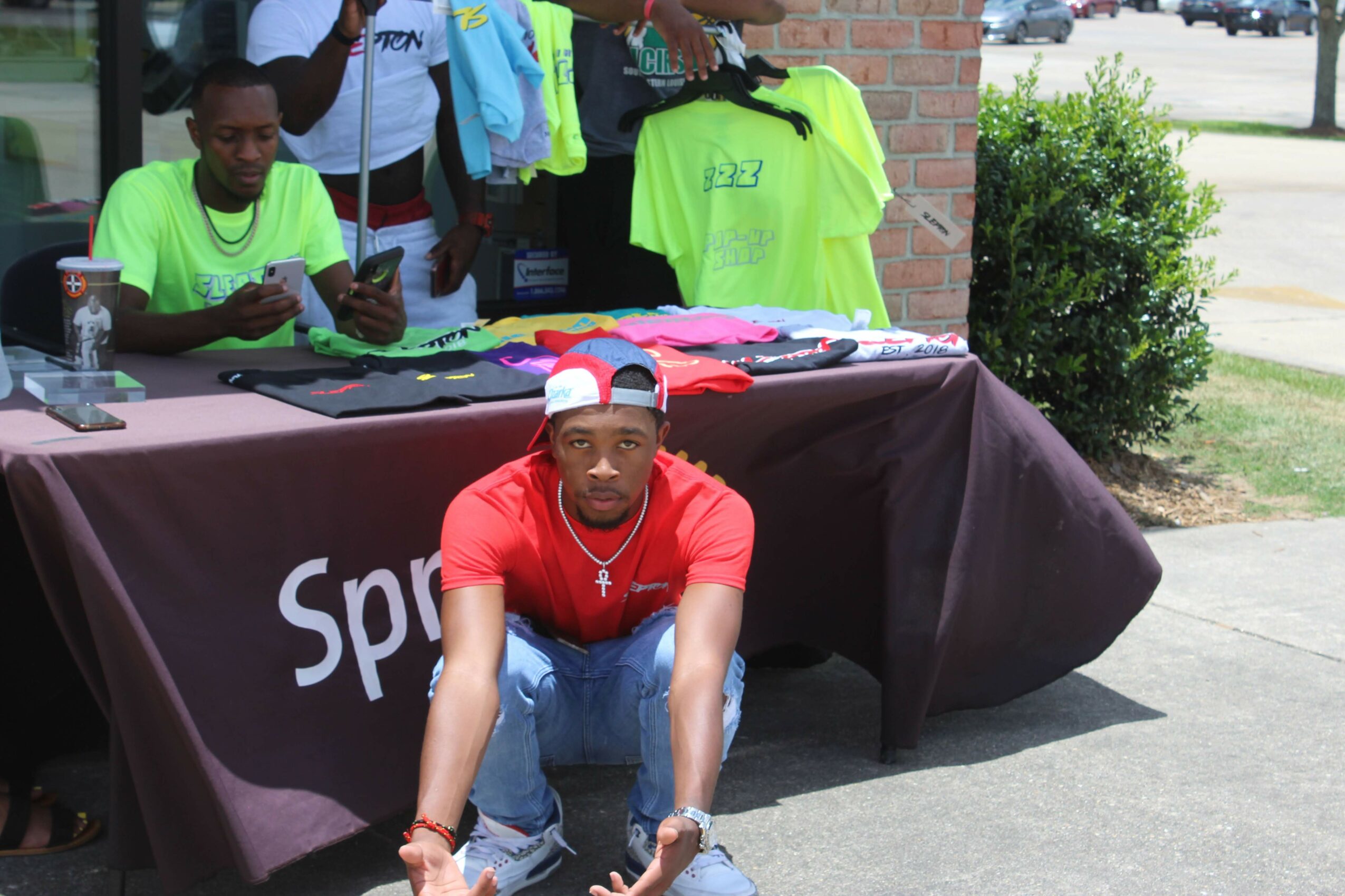 Young man in red shirt and cap sitting in front of a table with merchandise, people behind the table, outdoors.