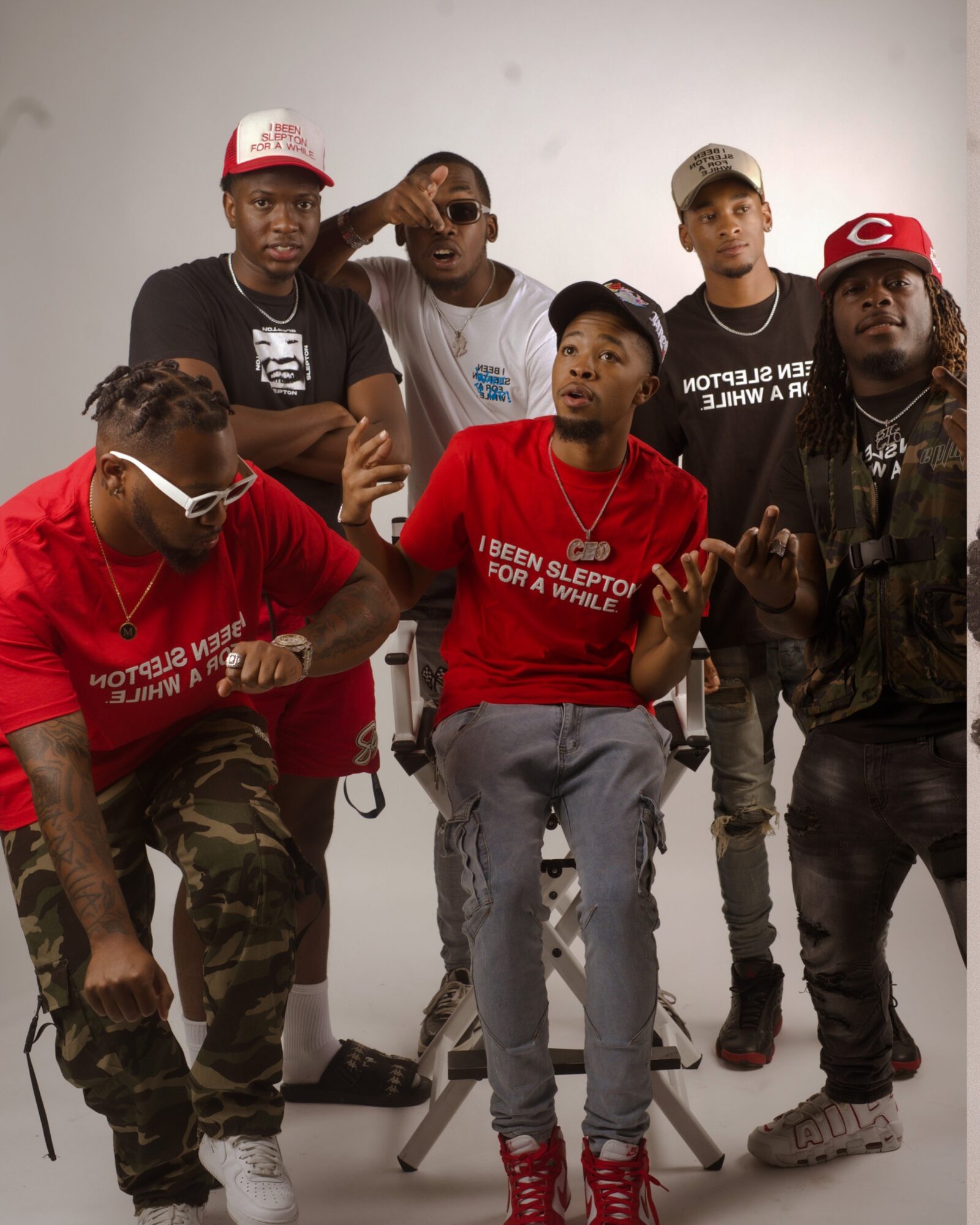 Group of six young men posing together, some making gestures, in casual clothing and hats, against a plain background.
