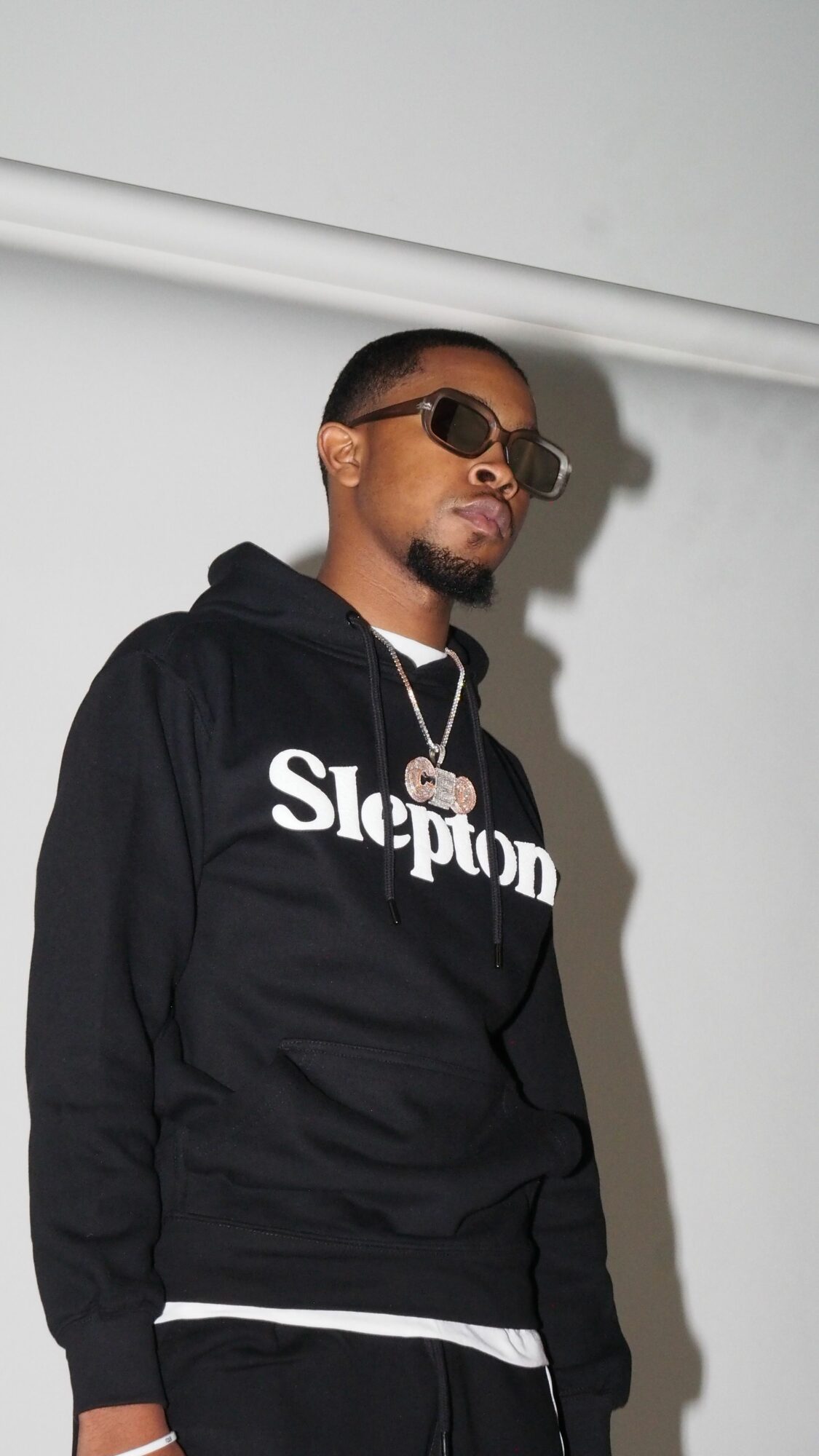 Person wearing sunglasses and a black hoodie with white text, standing against a plain wall.