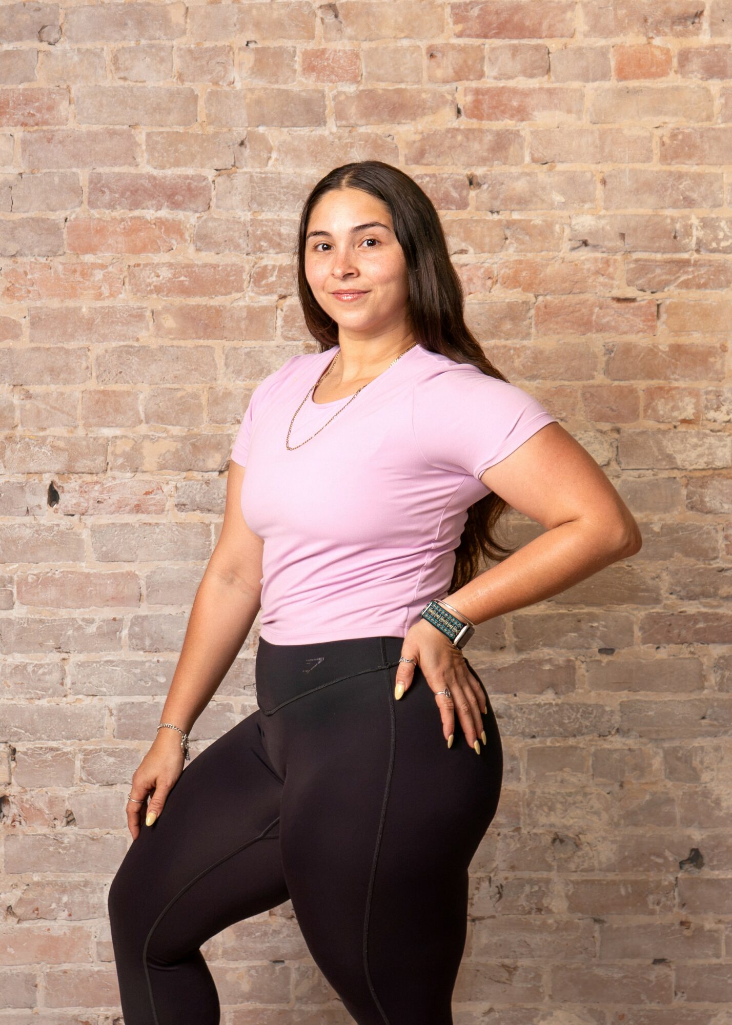 Young woman with long dark hair in a pink t-shirt and black pants standing against a brick wall, smiling, with one hand on her hip.