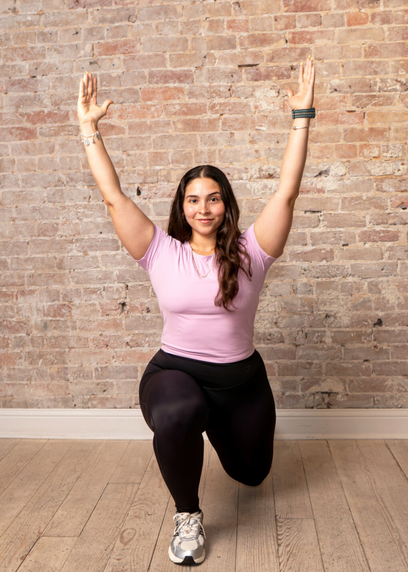 Woman in pink shirt and black leggings kneeling with arms raised in front of brick wall.