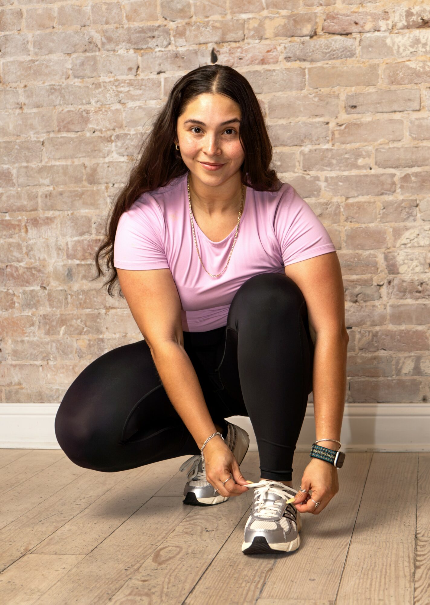 Young woman crouching, tying her shoe, wearing a pink shirt and black leggings, against a brick wall background.