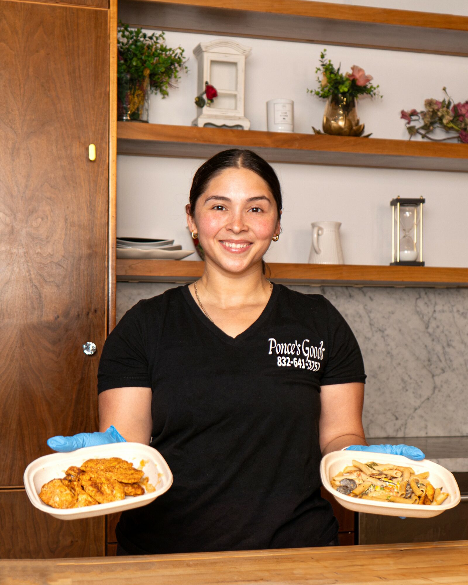 Smiling woman in black shirt holding plates of food in a kitchen with shelves and decorations behind her.