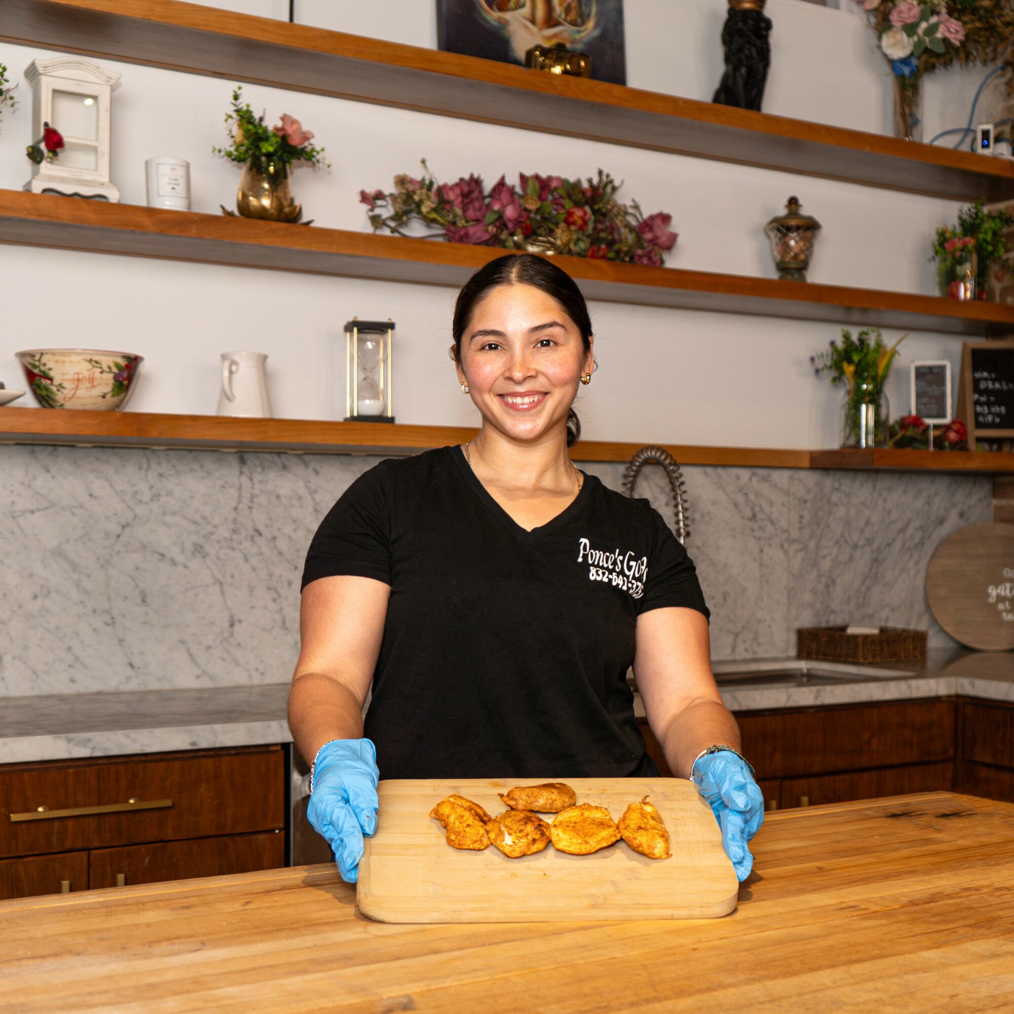 Smiling woman in black shirt and blue gloves holding a wooden tray with cooked chicken pieces in a kitchen with shelves and decorative items