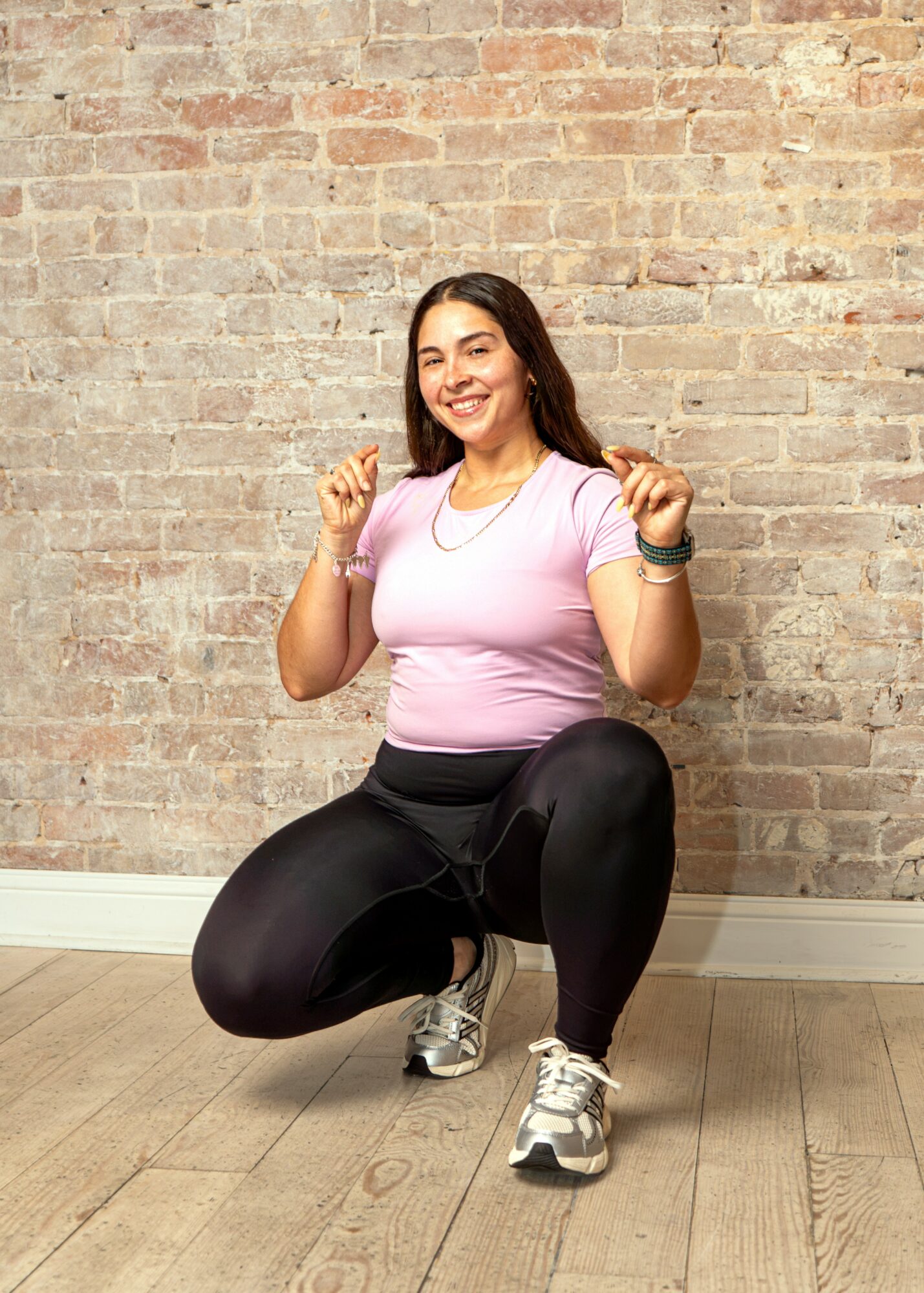 Smiling woman in pink shirt and black leggings crouches with fists raised against a brick wall.