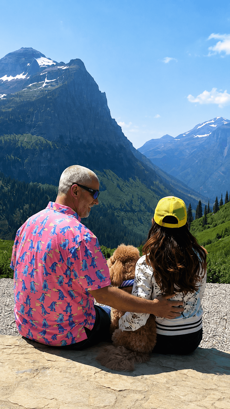 Two people and a dog sitting on a rock with mountains and blue sky in the background.