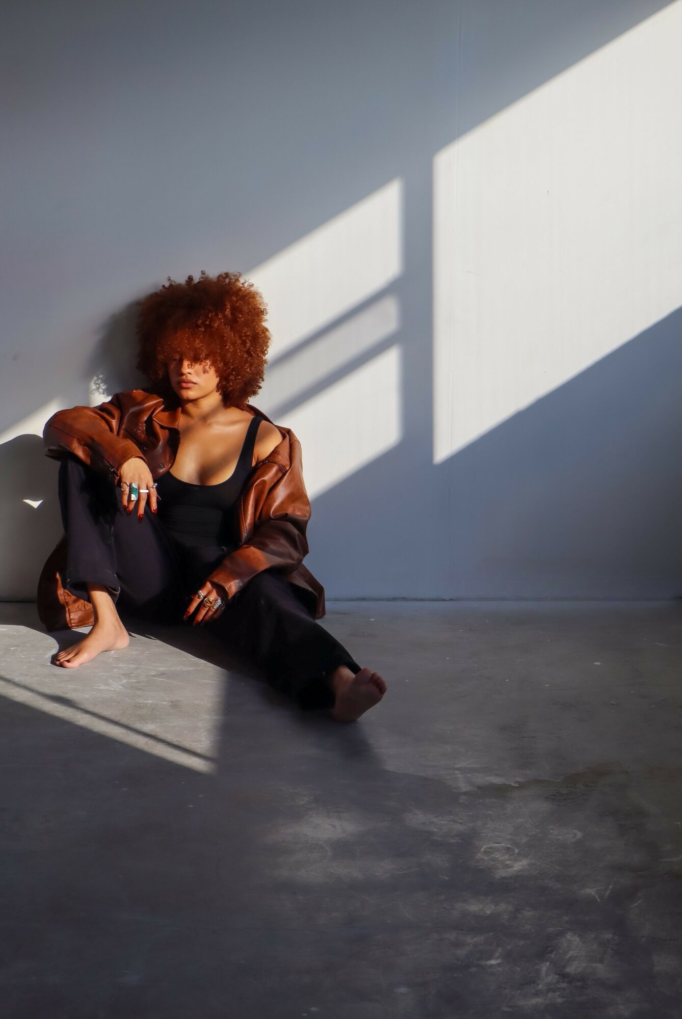 Person with curly hair sitting on the floor against a wall, with sunlight and shadow patterns on the wall.