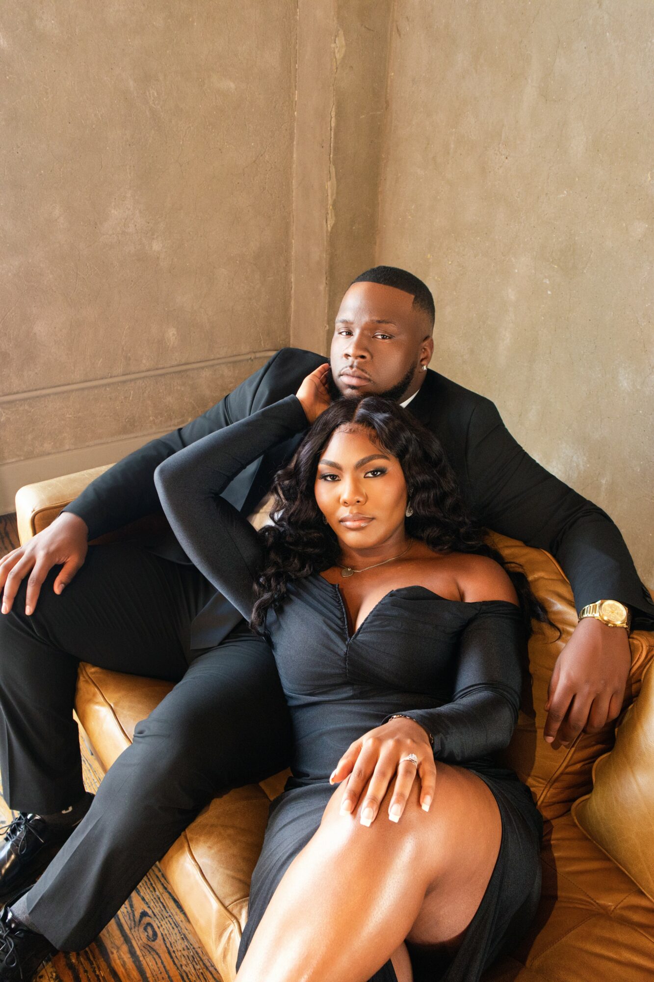 A man and woman sit on a yellow couch, with the woman in front and the man behind her, both dressed in black.