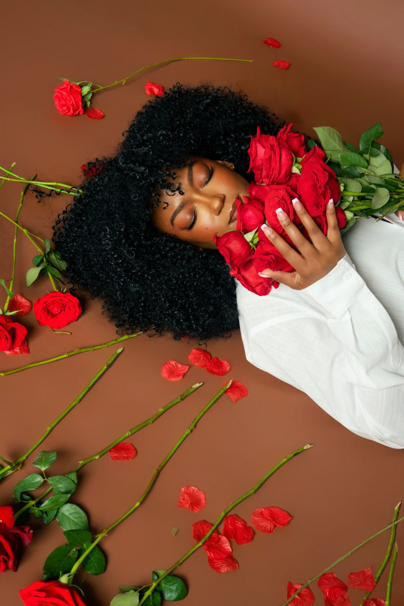 Woman with curly hair lying on brown surface, holding red roses, surrounded by scattered roses and petals, eyes closed, white shirt.