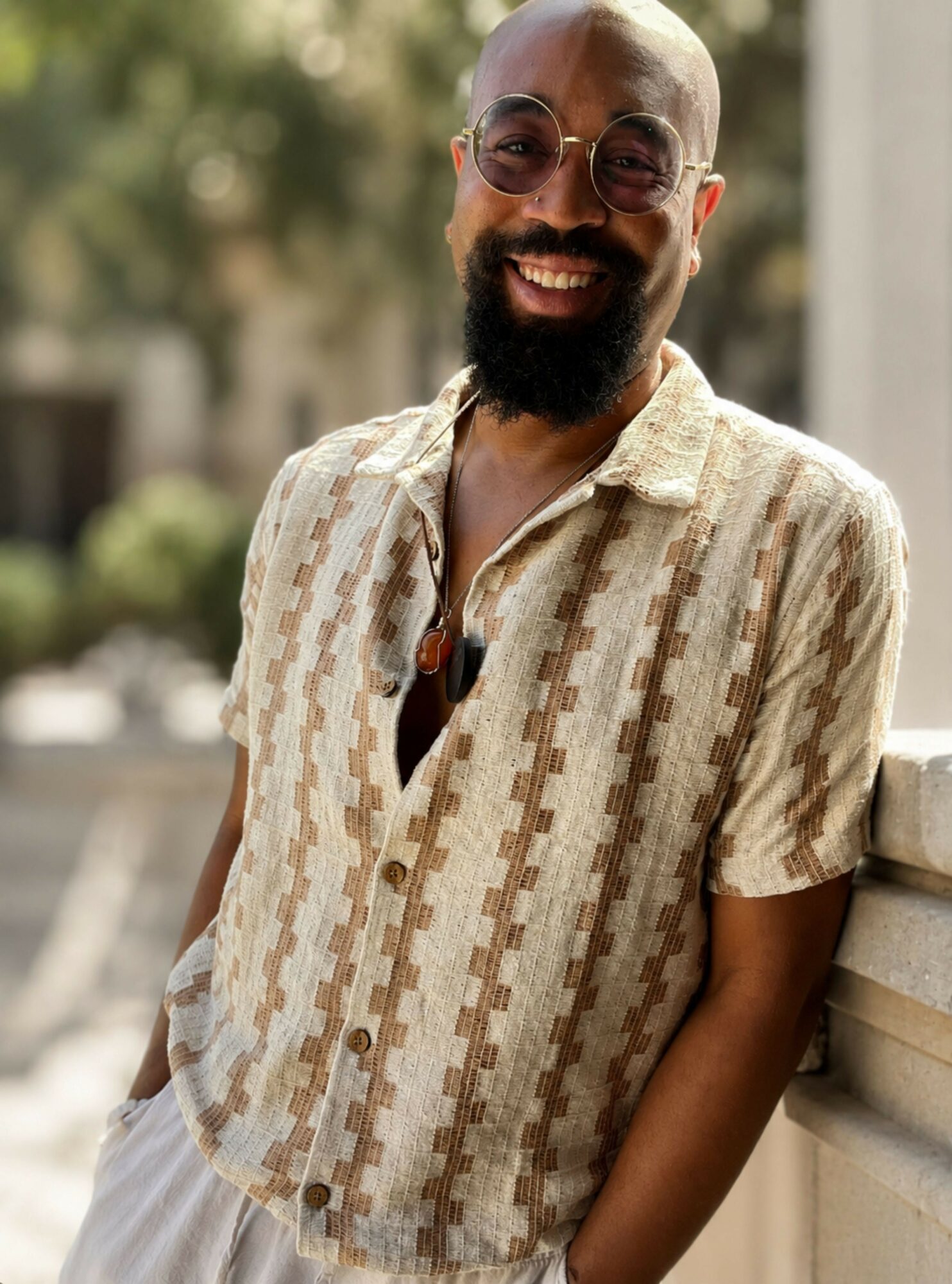Smiling man with glasses and a beard wearing a patterned short-sleeve shirt outdoors, leaning on a railing.