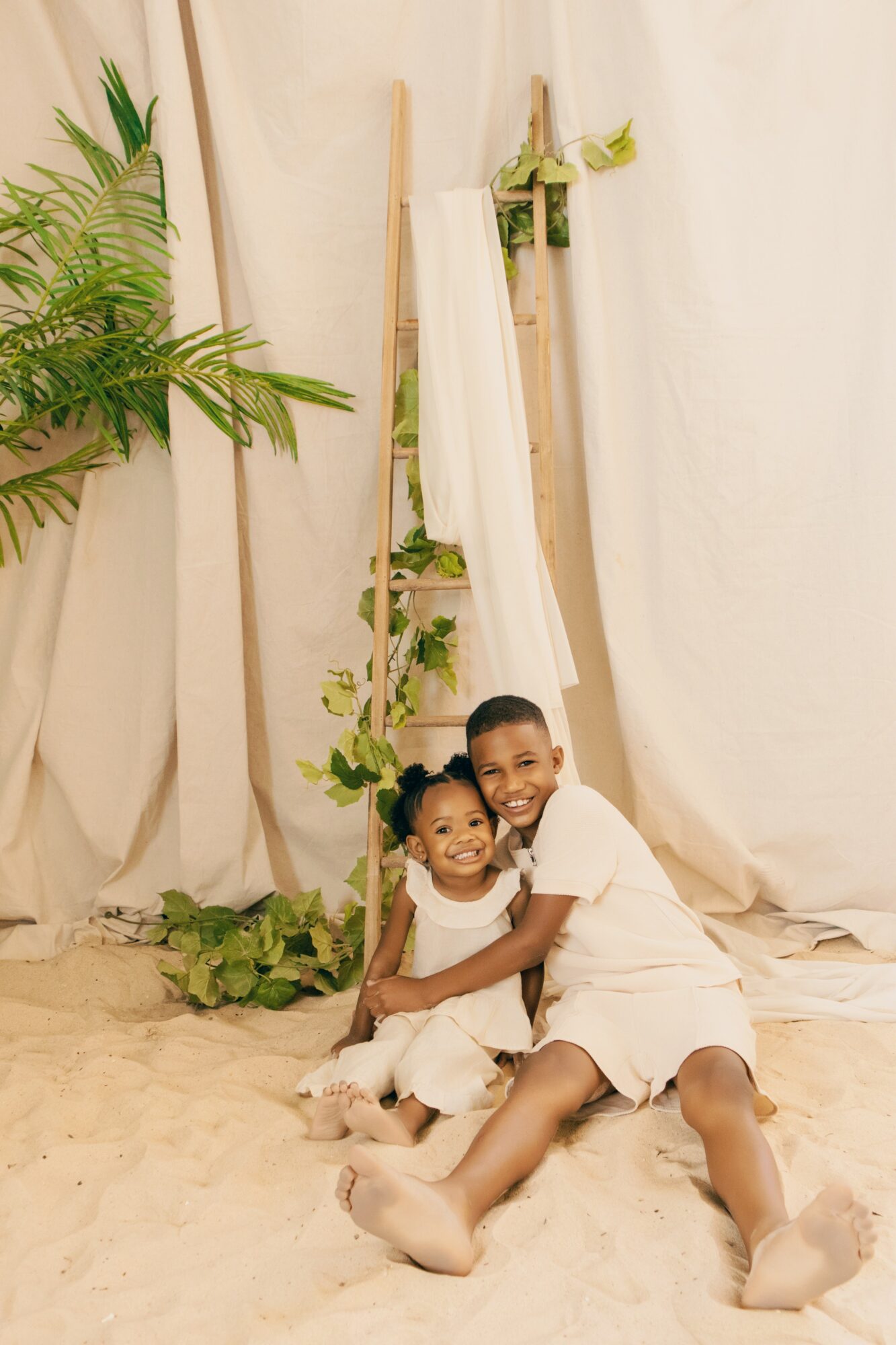 Two children sitting on sand, smiling, with a white fabric backdrop and green plants behind them.