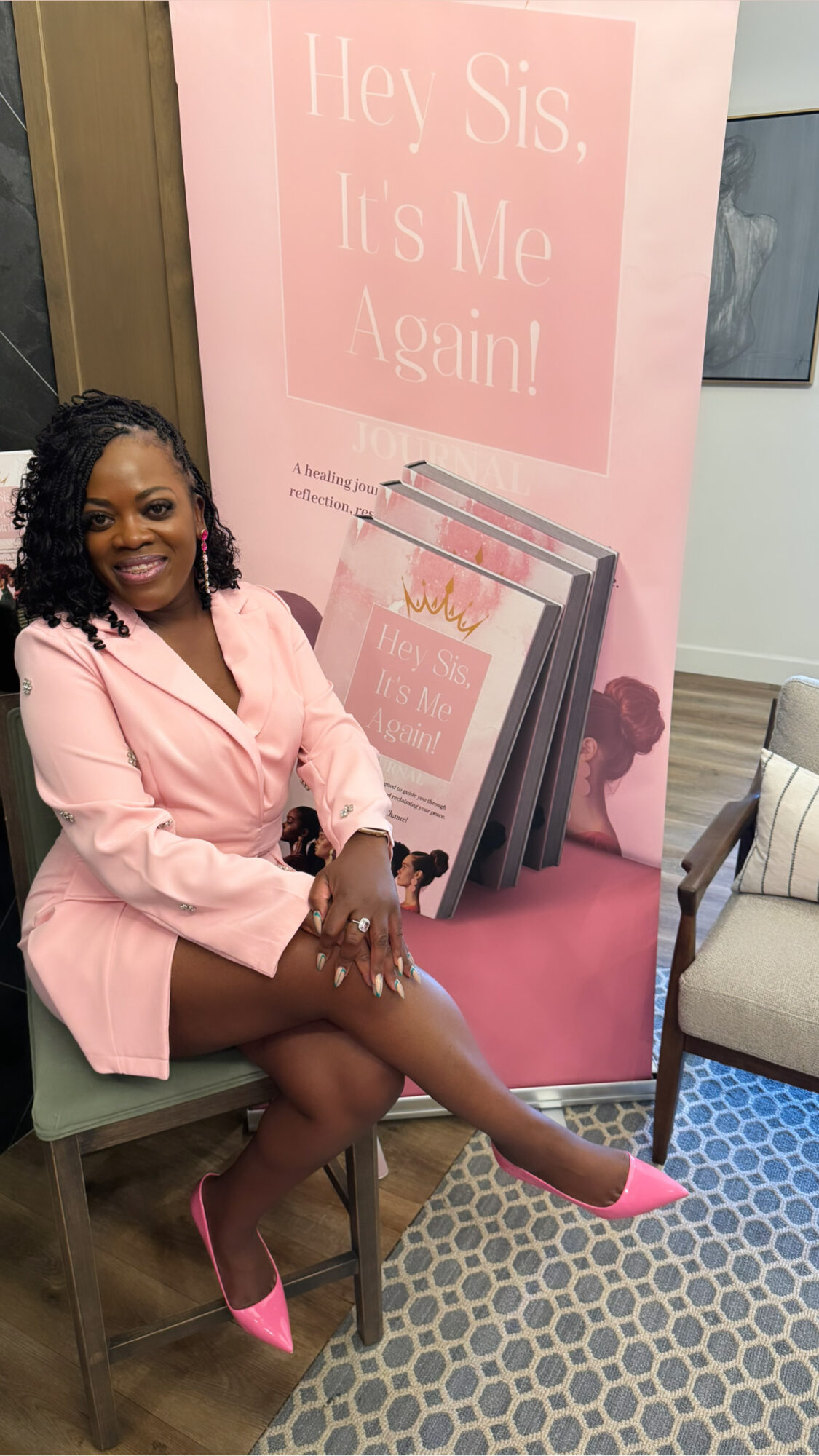 Woman in pink blazer and shoes sitting on a chair, smiling, with a pink book display behind her.