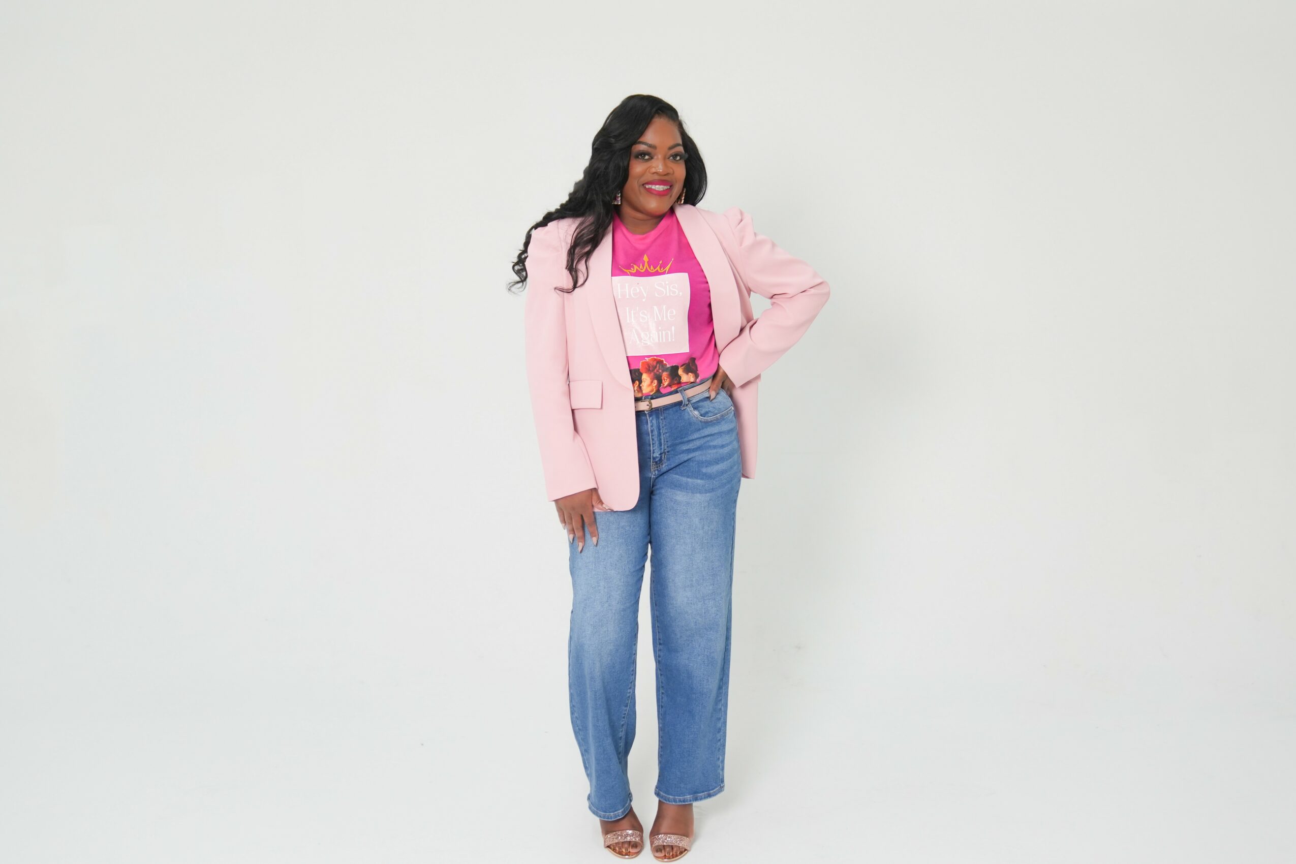 Woman with long dark hair wearing a pink blazer, pink top, and blue jeans, smiling and standing with one hand on her hip.