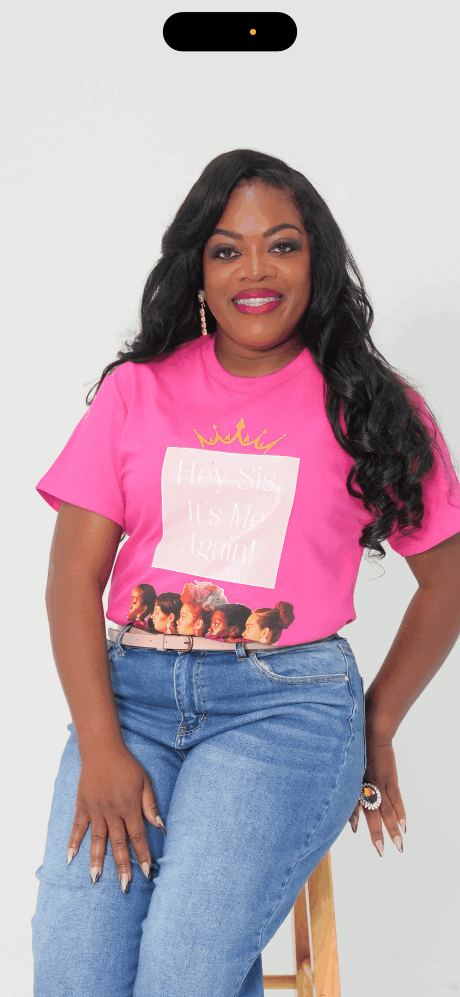 Woman with long black hair wearing a pink T-shirt and blue jeans, smiling, standing against a plain background.