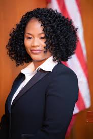 Woman with curly hair wearing a dark blazer and white shirt, standing in front of an American flag.