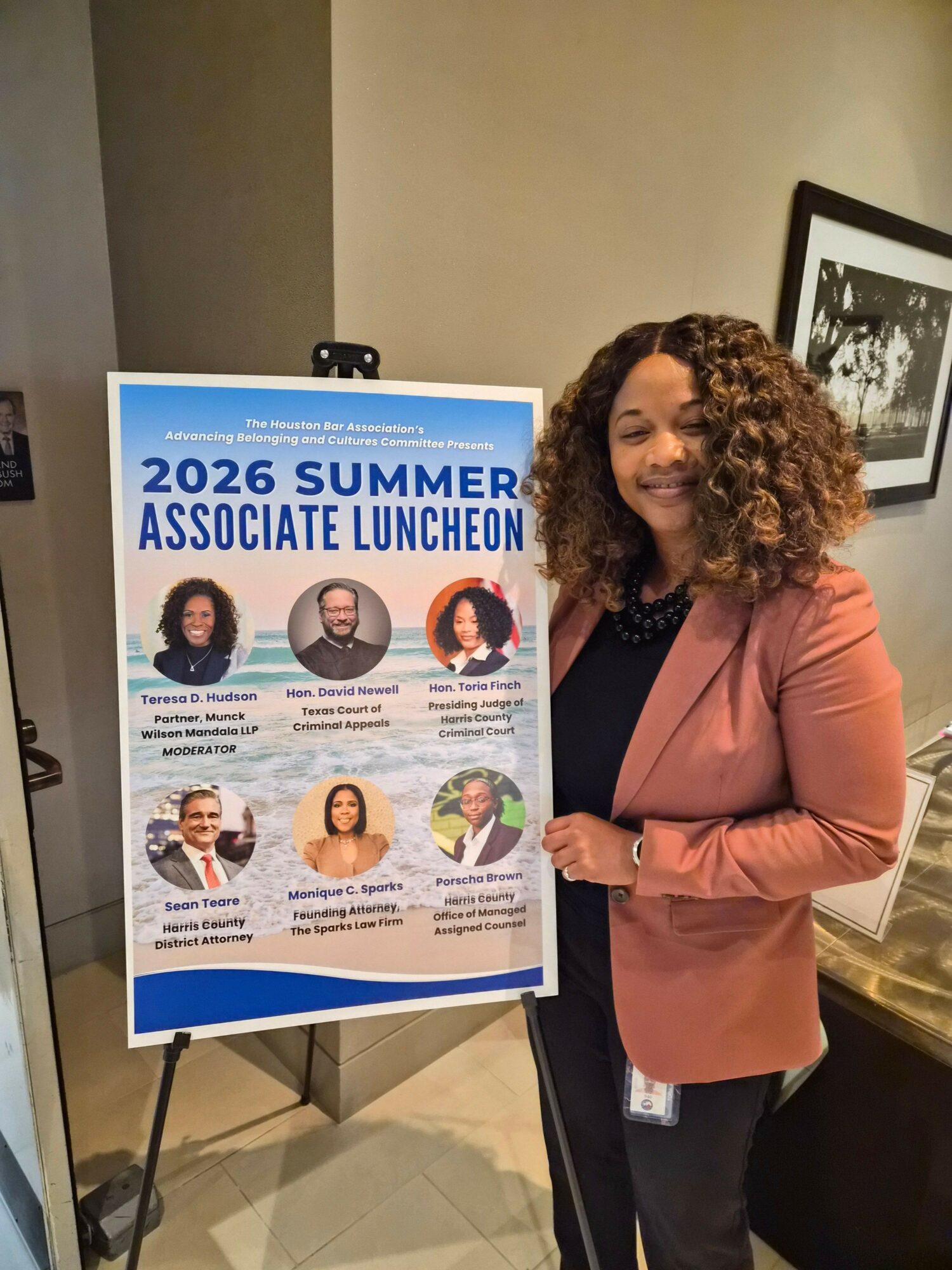 Woman with curly hair in pink blazer standing next to a poster about summer activities, indoors, with framed picture on wall