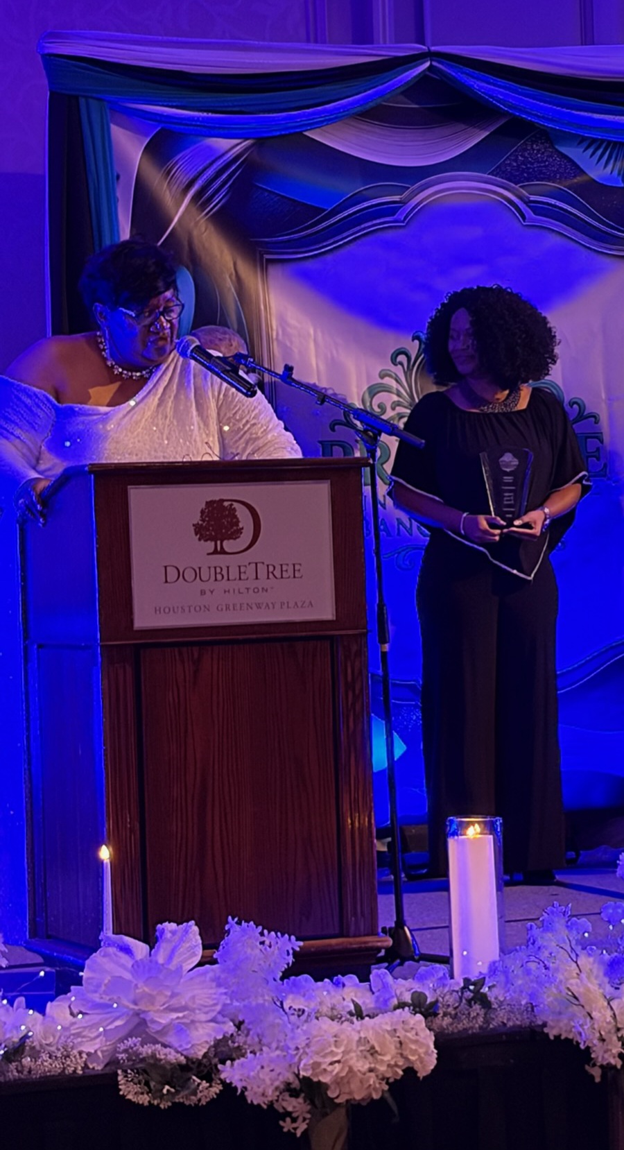 Two women stand at a podium with a sign reading 'DoubleTree,' surrounded by flowers and candles, under blue lighting.