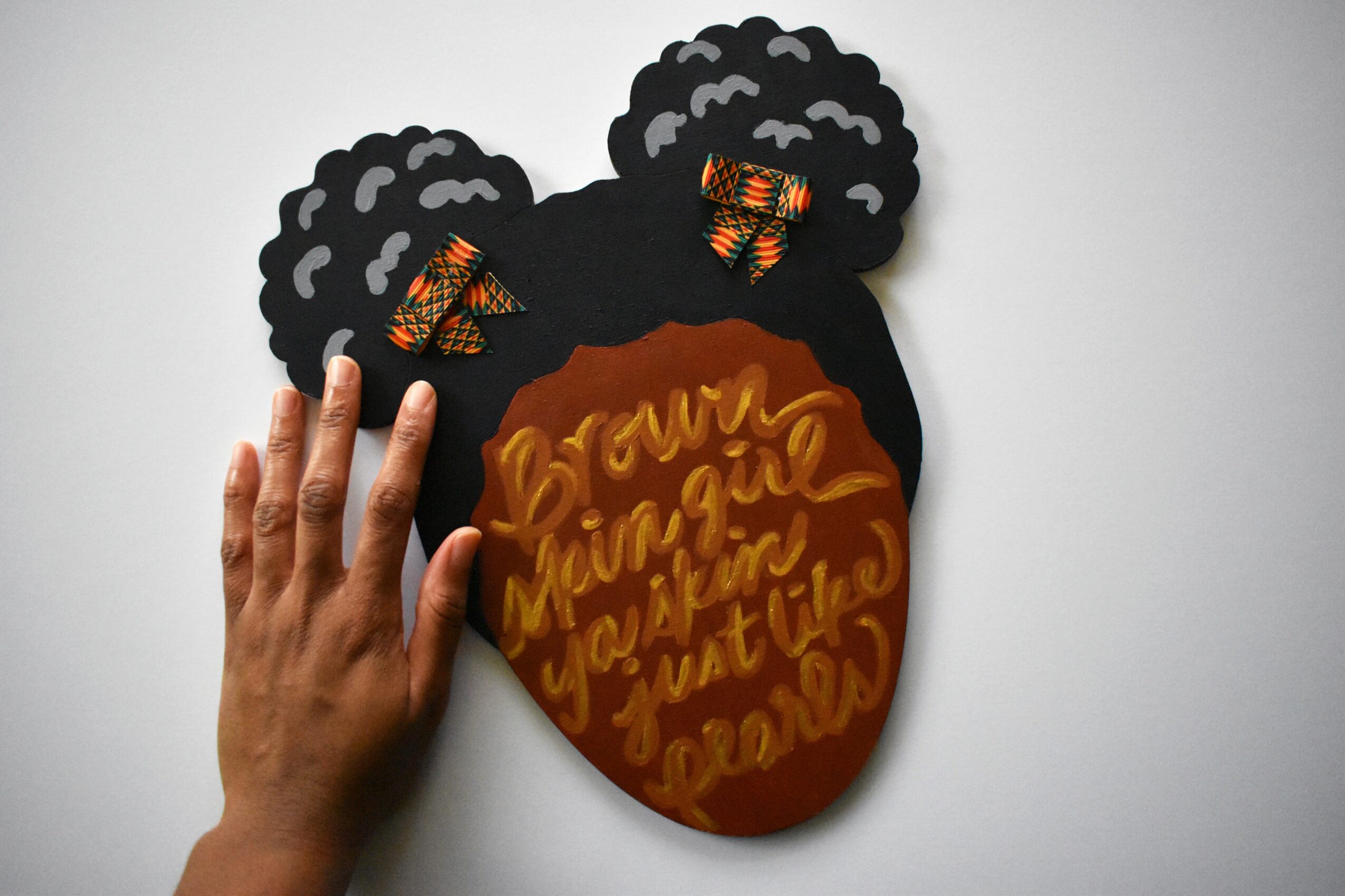 Hand holding a black paper cutout of a head with curly hair, orange face with handwritten text, against a white background.