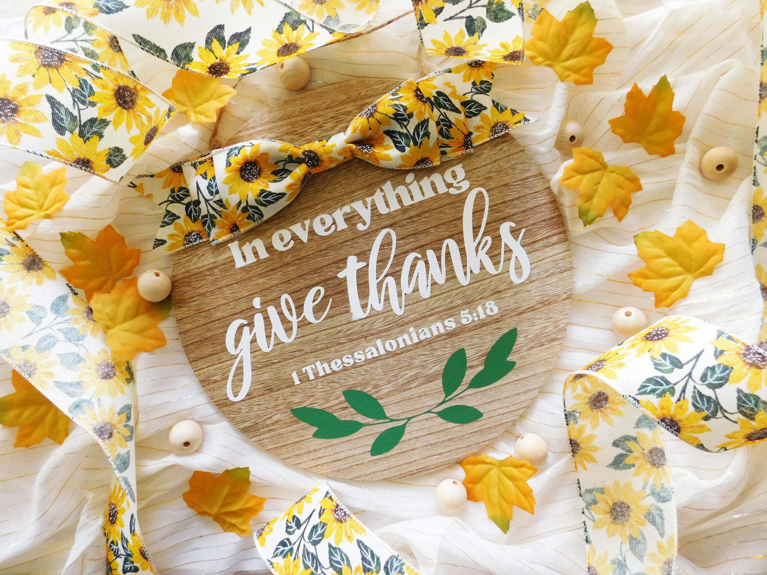 Wooden sign with white text, surrounded by yellow leaves, fabric with sunflower pattern, and small white beads.