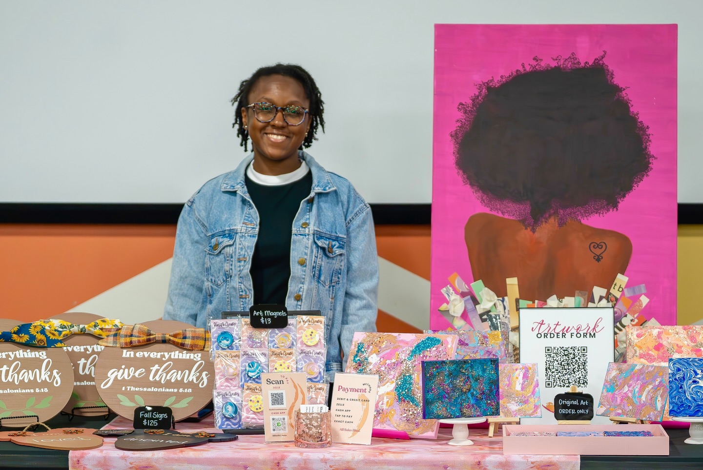 Smiling person standing behind a table with various gift items and a pink painting of a woman with curly hair.