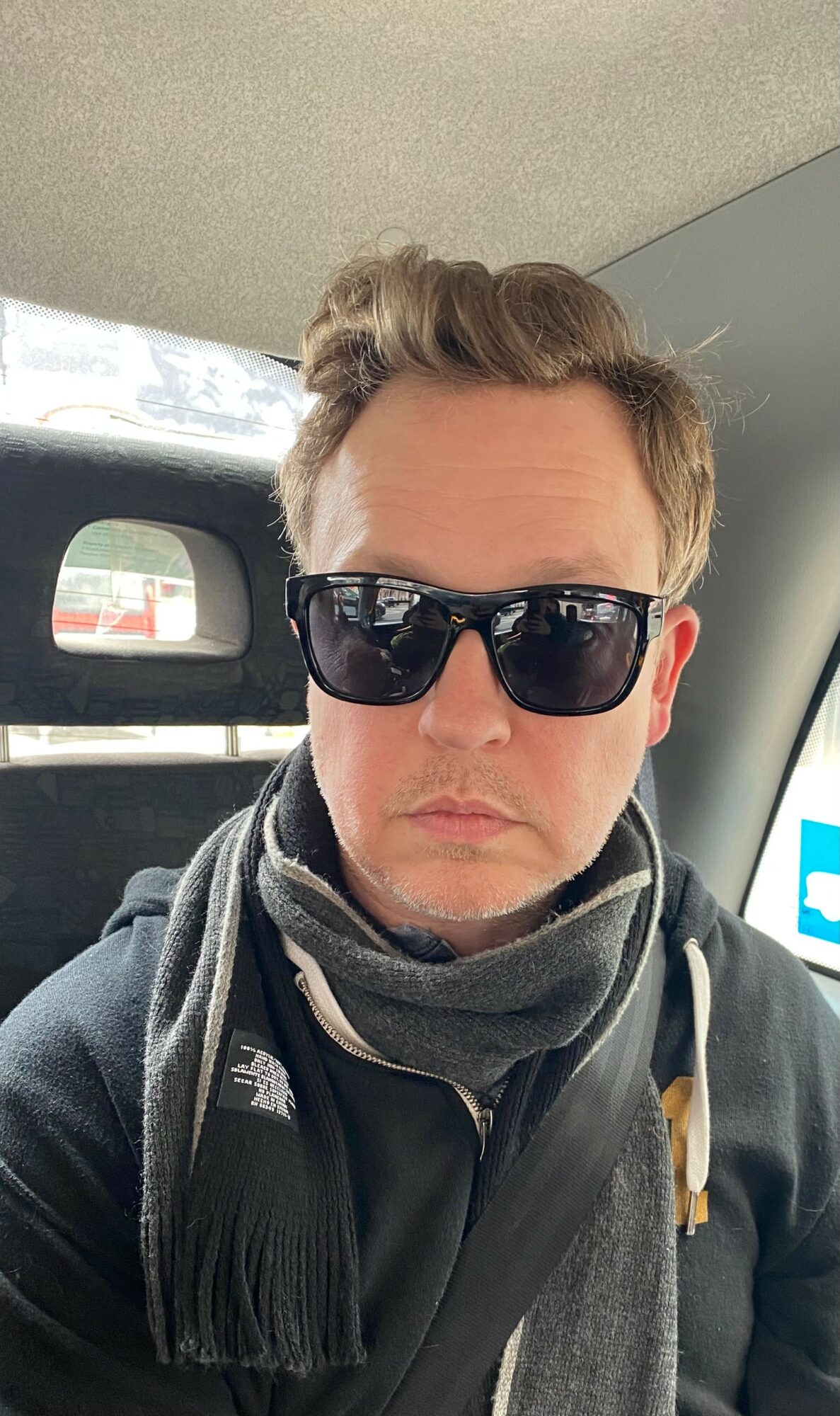 Person wearing sunglasses, scarf, and jacket inside vehicle, with a window showing a snowy landscape behind.
